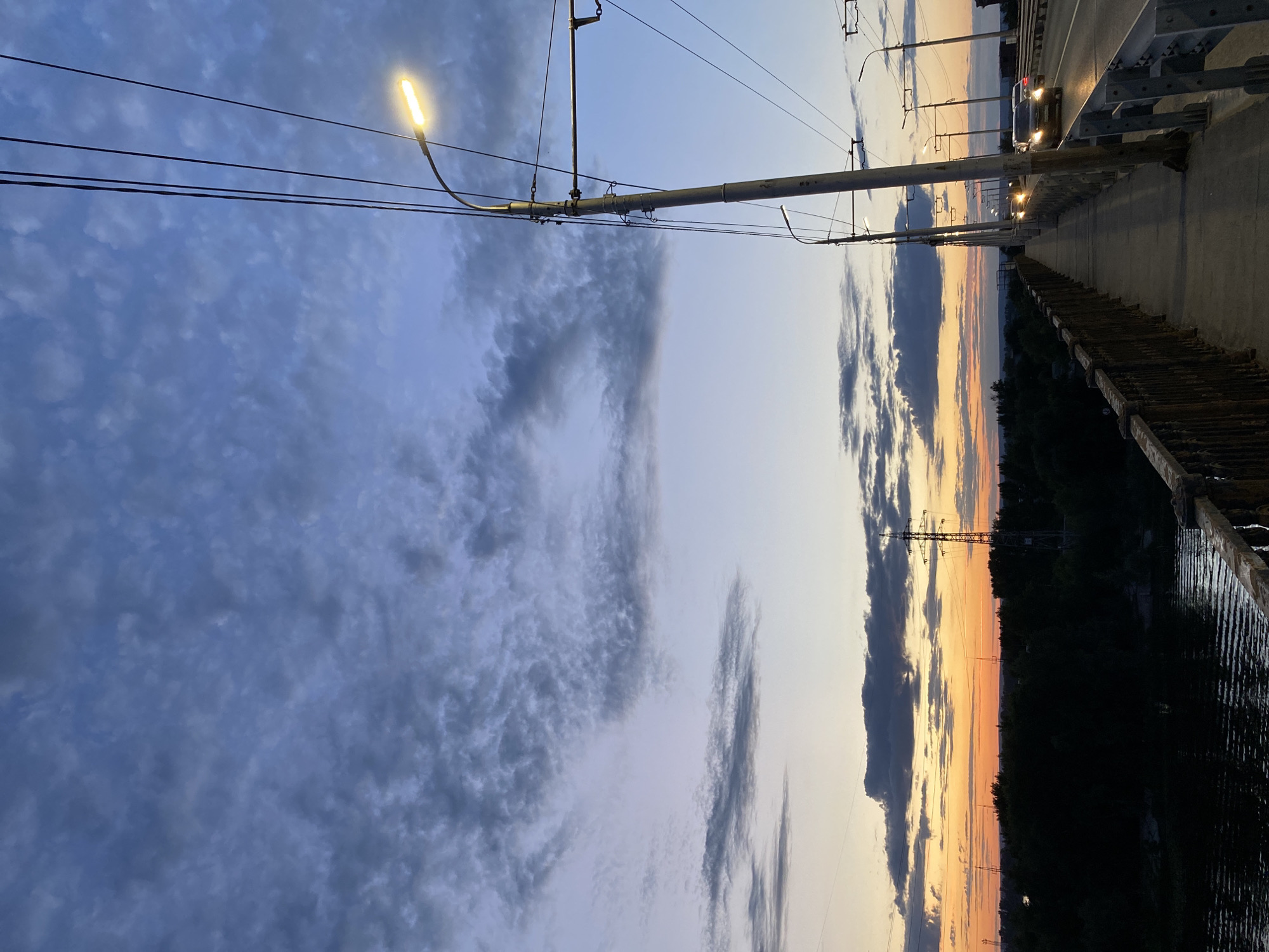 This image captures a view from a bridge over a body of water in Novokuybyshevsk, Russia, at dusk. The sky is filled with dramatic clouds, transitioning from a deep blue at the top to hues of orange and yellow near the horizon, indicating the setting sun. Power lines and streetlights stretch across the scene, with one streetlight illuminated. A road runs parallel to the pedestrian walkway on the bridge, and several cars are visible on the road, their headlights on. The landscape beyond the bridge is dark and features a dense line of trees silhouetted against the twilight sky, with power transmission towers also present in the distance. The pedestrian walkway has a metal railing and a concrete surface. The water below reflects the dim light.