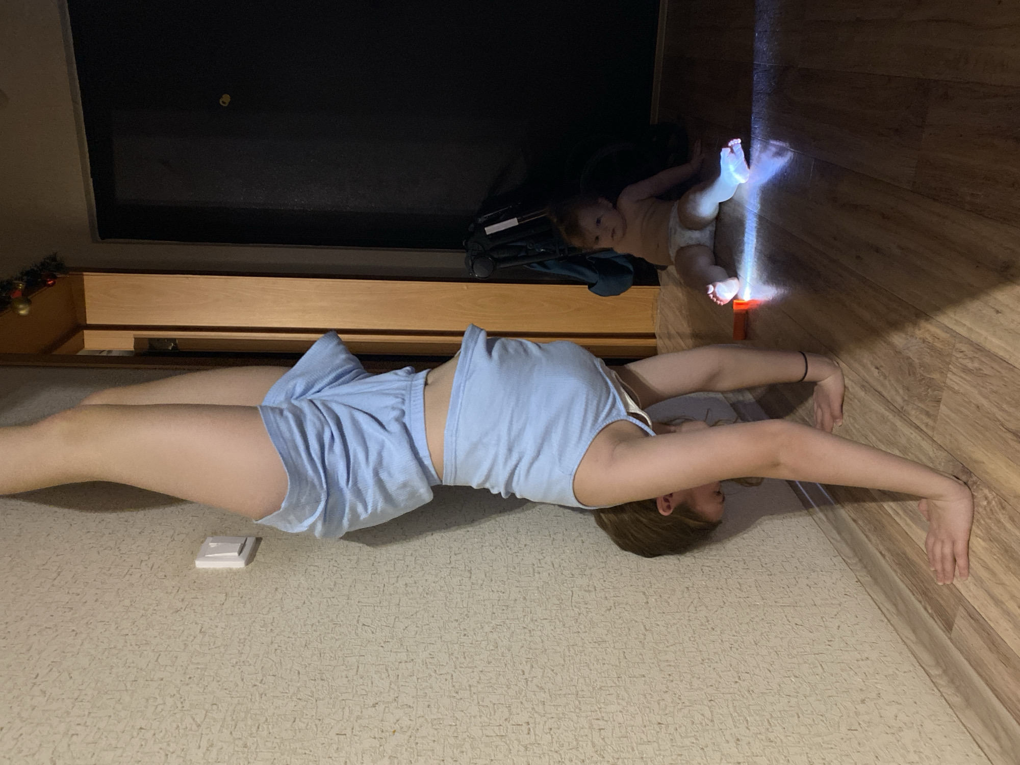 A woman in light blue loungewear stretches horizontally on a beige carpeted floor, with her head towards the lower right. Her arms are extended overhead, reaching towards a wooden-paneled wall. A baby in a diaper lies on a wooden floor near the top right, illuminated by a beam of light. Behind the baby are indistinct objects including a black object resembling a stroller. Above the woman's head and to the left is a shelf and dark rectangular object. A small, square white object is on the carpet near the woman's midsection.