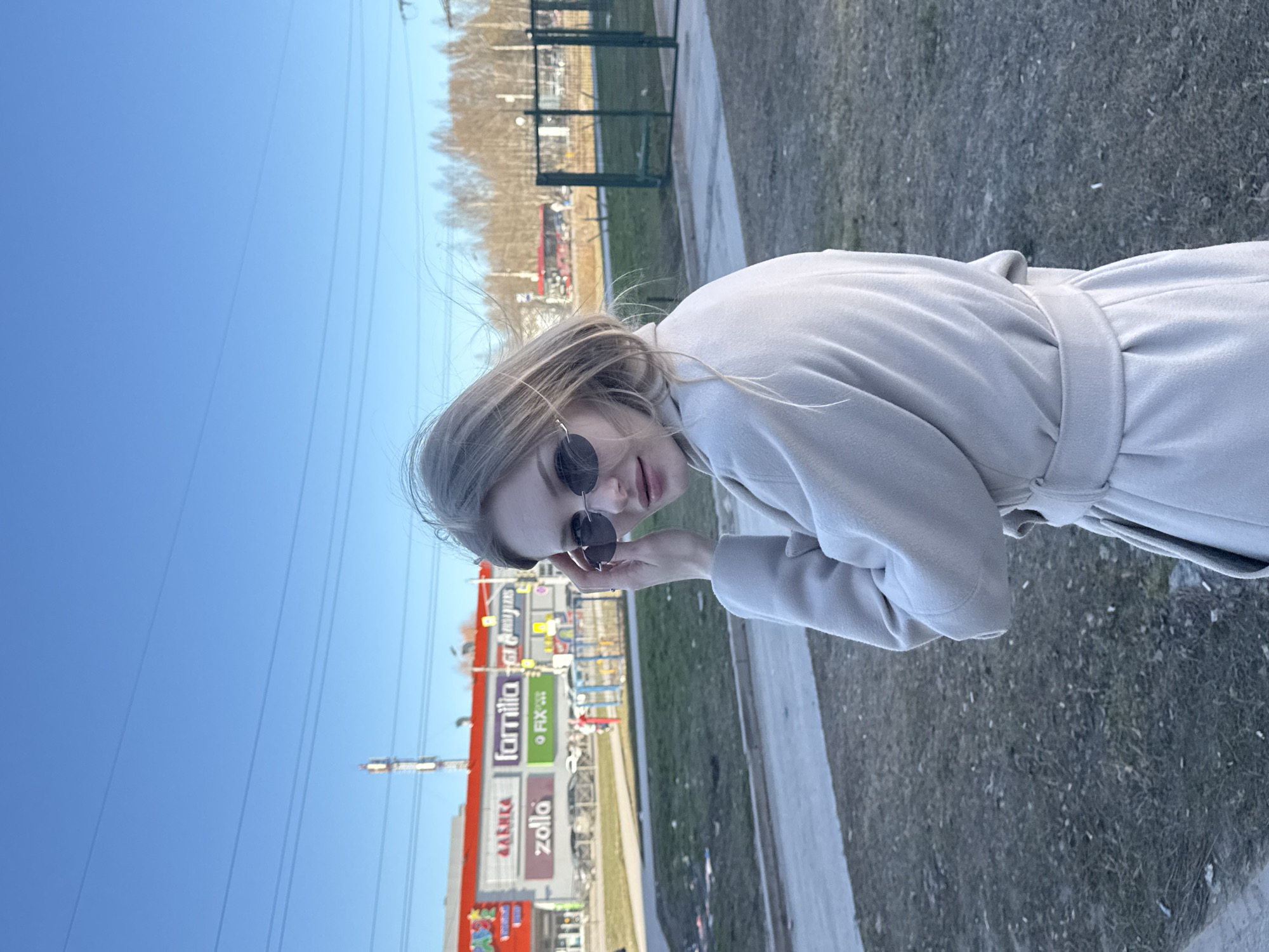 A fair-skinned woman with blond hair and sunglasses stands outdoors, wearing a light-colored coat with a tied belt. She looks toward the camera and is holding her sunglasses near her eye. The background shows a shopping center with signs in Russian, including "Familia" and "Zolla", located in the city of Perm, Russia. Overhead utility lines cross the sky. The ground is covered in dormant vegetation and some pavement is visible.