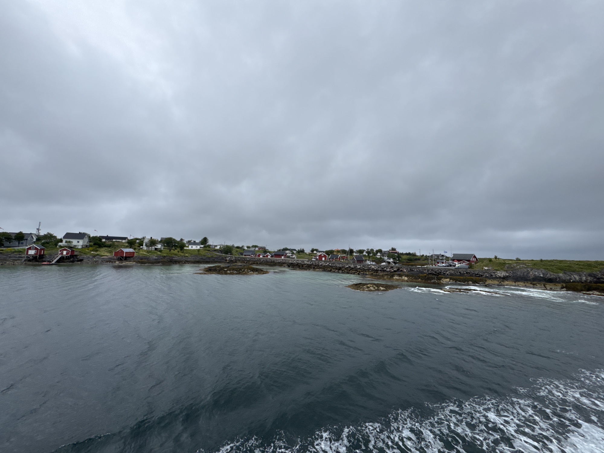 The image shows a view of the Sleneset area in Norway from the water. A small coastal village with several red and white houses is visible on the shore. The houses are situated on rocky terrain with some greenery. The sea in the foreground is dark and choppy, with white foam indicating motion, possibly from a boat. The sky is overcast with heavy gray clouds, suggesting a cloudy or possibly rainy day. The scene is natural and depicts a typical northern coastal landscape. There are no people visible in the image.