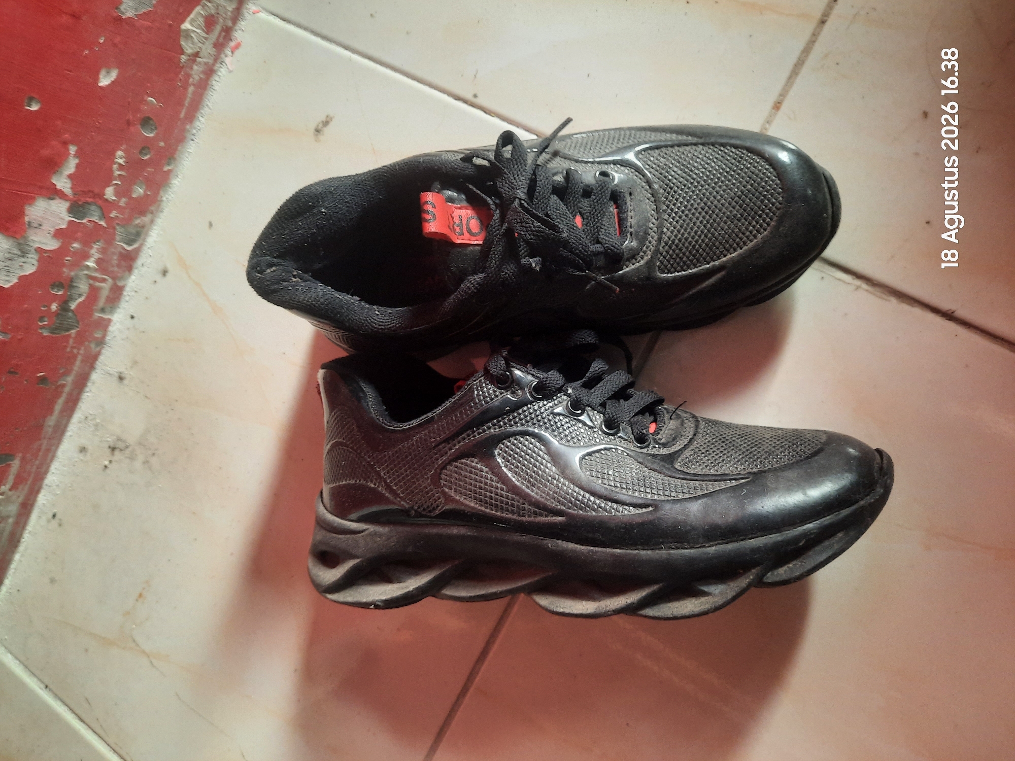 A pair of black athletic shoes are positioned on a tiled floor. One shoe is lying on its side, revealing its textured sole and mesh upper. The other shoe is upright, with black laces tied. A red tag with orange lettering is visible inside the upright shoe. The background shows a red, textured wall and a tiled floor with visible grout lines. A timestamp, "18 Agustus 2026 16.38," is present in the lower right corner, indicating the date and time the photograph was taken. The location is identified as Sukokerto, Indonesia.
