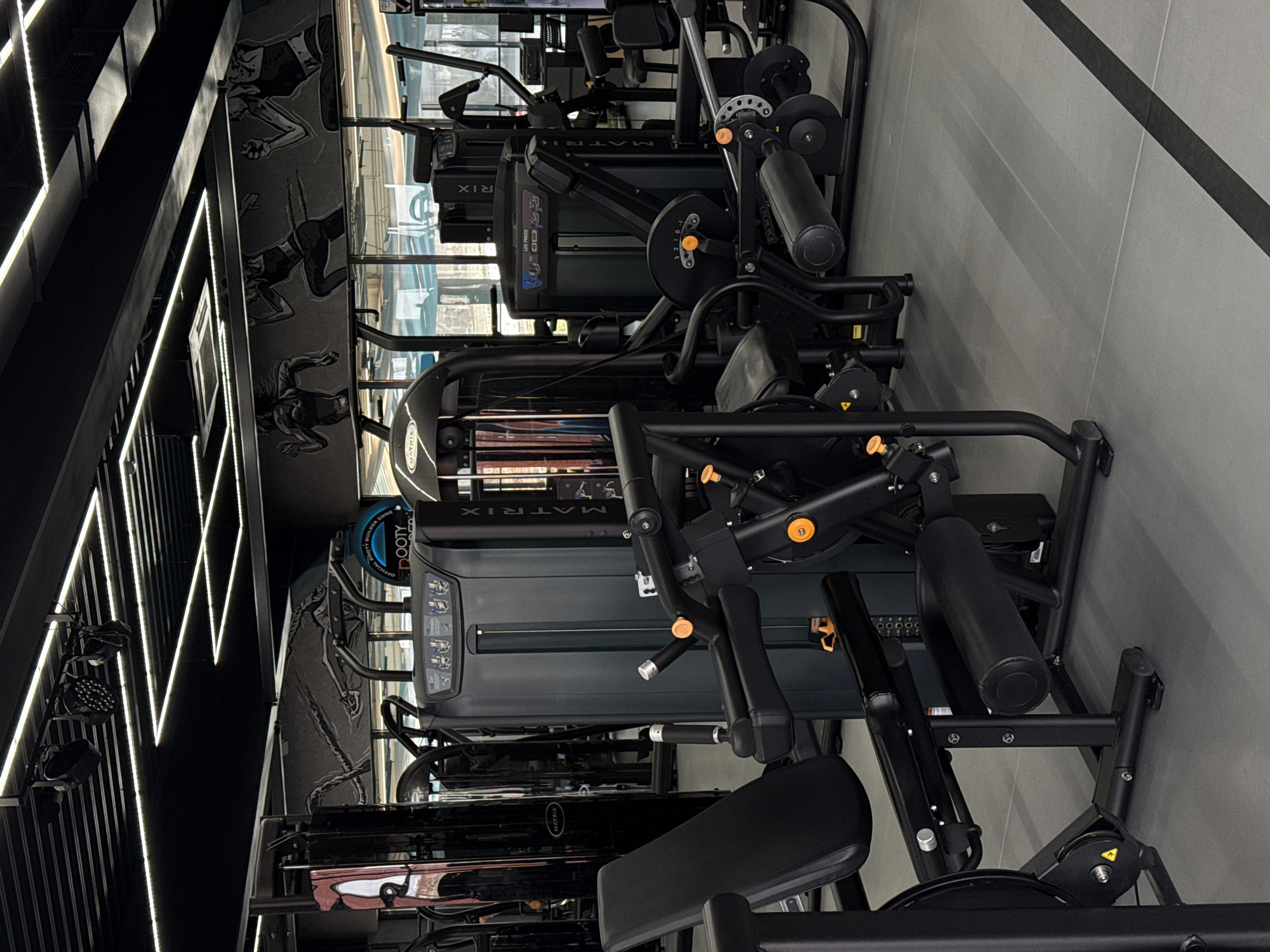The image depicts a gym interior, likely located in a commercial space. Several pieces of Matrix-branded gym equipment are arranged in rows, occupying most of the frame. The equipment includes leg press machines and other weight-training apparatus. The gym's interior design features black walls with vertical LED light strips and graphic murals of athletes. The floor is gray, and a black stripe runs diagonally along one side. The environment appears well-lit, clean, and organized. Given the prompt specifying Votkinsk, Russia, this gym may be located within that city.