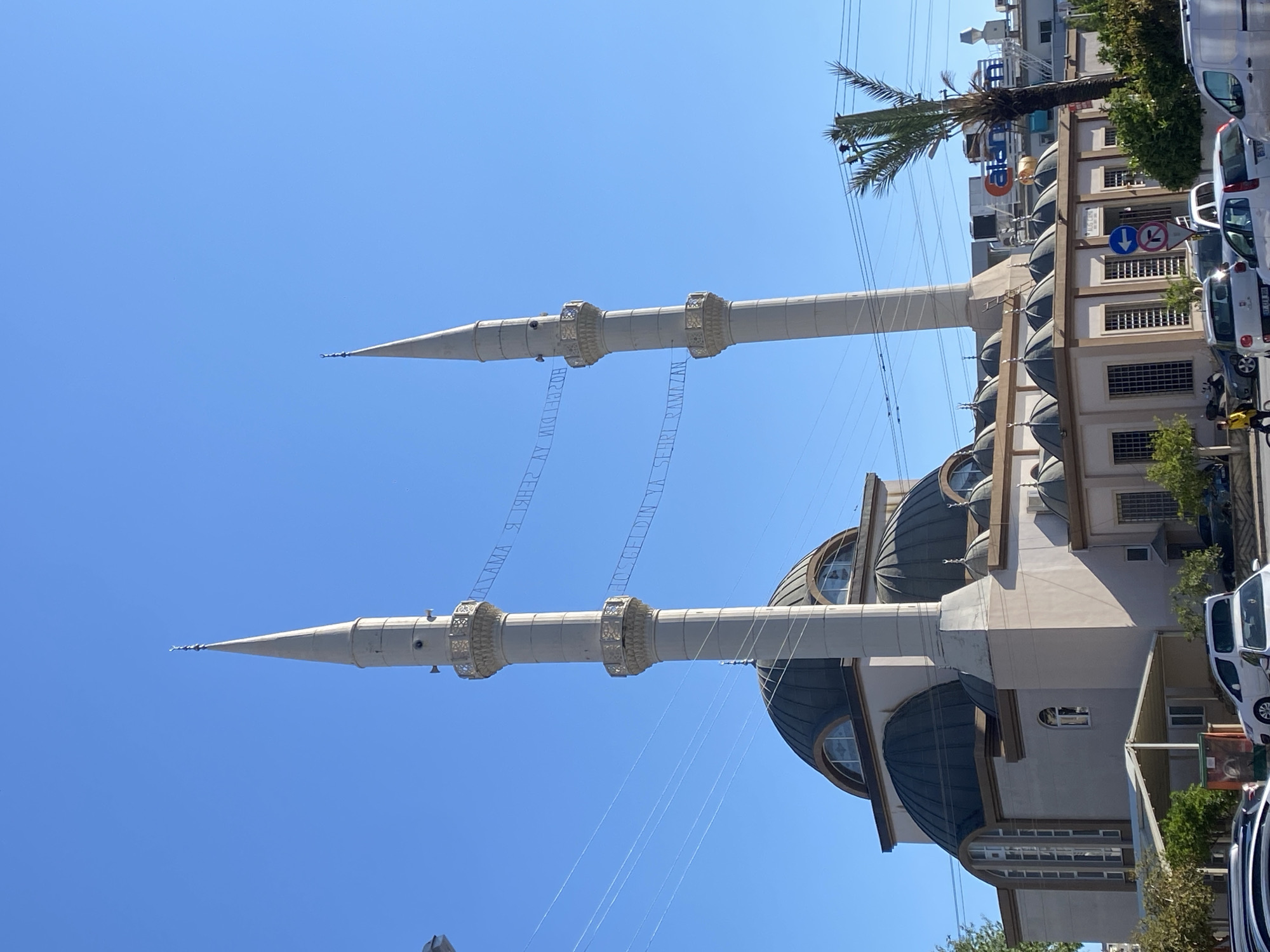 The image, taken in Alanya, Turkey, captures a mosque under a clear, bright blue sky. The mosque features a light-colored facade with multiple dark, bulbous domes. Two prominent, slender minarets, each with two balconies and a pointed spire, frame the upper part of the mosque.
Between the two minarets, a large banner is strung, displaying text. When properly oriented, the text reads "HOŞ GELDİN AY" on the left and "ŞEHRİ RAMAZAN" on the right. This translates to "Welcome, O Month/City of Ramadan," indicating the photograph was taken during the Islamic holy month of Ramadan.
To the right of the mosque, a street scene unfolds. Several cars, including white vans and sedans, are parked along the street. A tall palm tree with lush green fronds stands near the mosque, providing some shade. Other buildings are visible in the background, including a multi-story structure with numerous windows. A blue circular road sign with white arrows (one pointing down, one pointing up with a red diagonal line, likely indicating a one-way street or restricted access) is also visible. The bright, cloudless sky suggests a sunny day. No specific activity or people are distinctly visible.