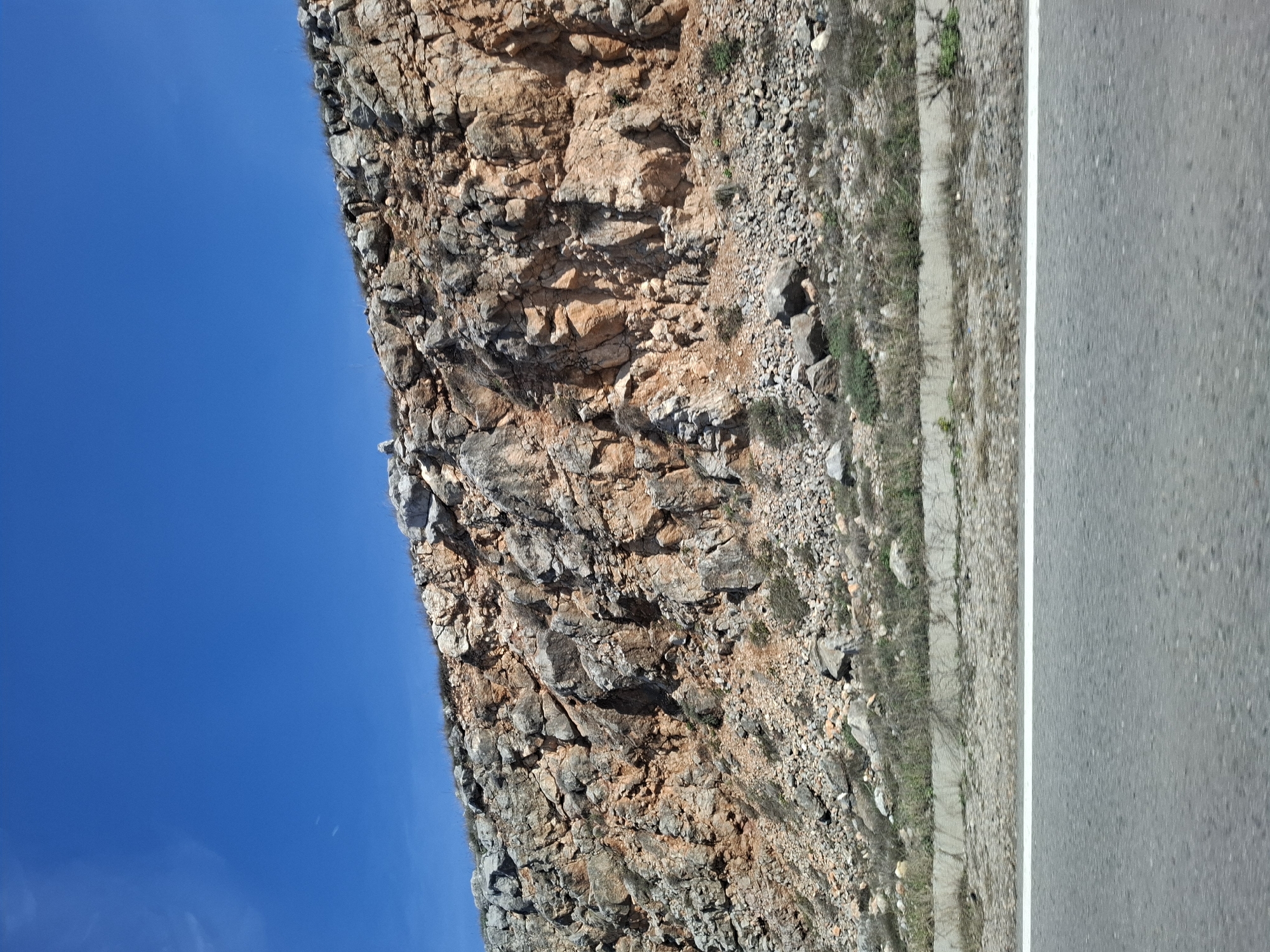 The image depicts a rocky cliff face adjacent to a road, with a clear blue sky visible above. The cliff is composed of various sized rocks and stones in shades of grey and brown. There are small patches of vegetation growing on the cliff face and along the roadside. The road is paved and marked with a white line. The location is Bogyovtsi, Bulgaria, based on the available information. The scene contains no people, subjects, objects, actions, interactions.