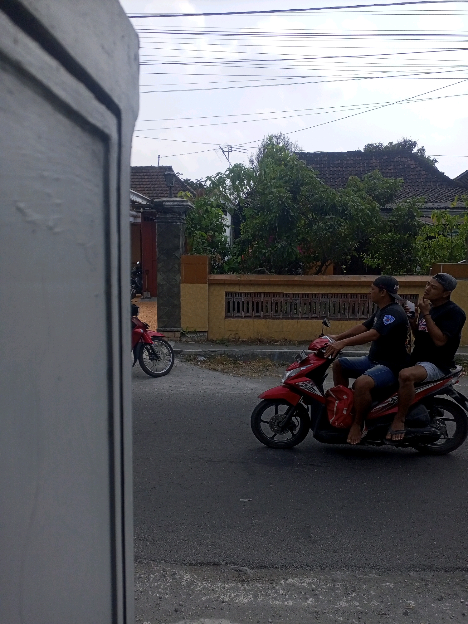 Two men ride on a red motorcycle on a paved road in front of a yellow and brown fence. The man in front is wearing a black t-shirt and blue shorts and is operating the motorcycle. The man behind him is wearing a black t-shirt and light blue shorts and is smoking. Both men are wearing baseball caps. The motorcycle has a red backpack attached to the back. Behind the fence are houses with tile roofs and trees. The sky is overcast. The image is framed by a large, grey, vertical structure on the left side, which appears to be a wall or pillar. The location is identified as Ngasem, Indonesia.
