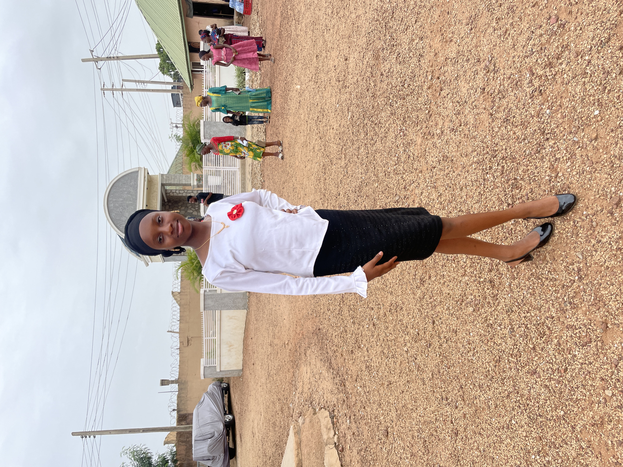 A young woman in a white blouse and black skirt stands on a gravel surface. She is looking at the camera with her right arm on her hip and her left arm at her side. In the background, other women are visible, wearing different colored dresses. A building with a green roof and a barbed wire fence are also visible. An overcast sky can be seen above. The location is Jos, Nigeria, as indicated by the gravel surfaces, architecture, and clothing of the subjects.