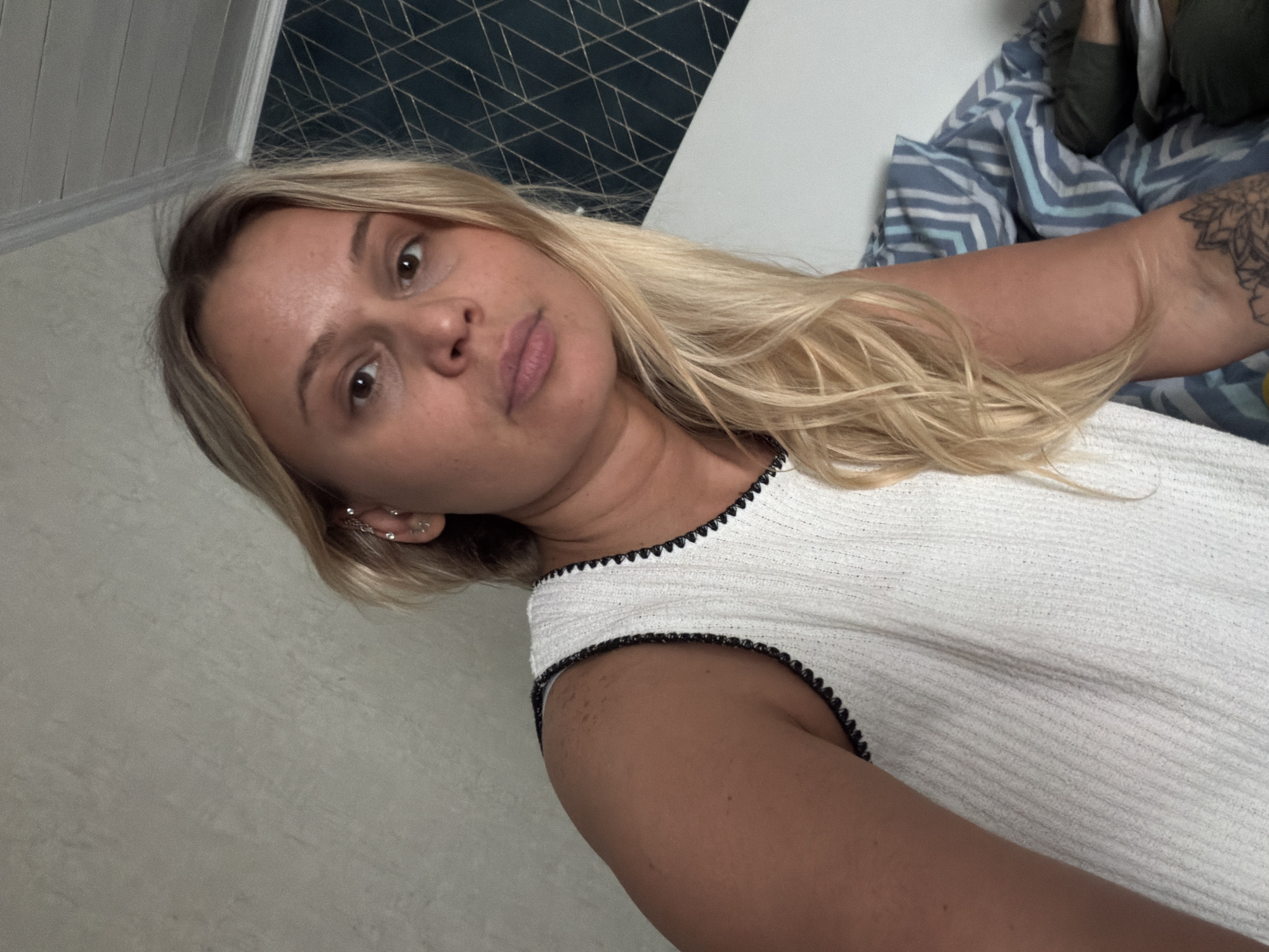 A blonde woman is depicted from a low-angle, close-up perspective. She wears a white top with black trim around the neckline and armhole. Her left arm is visible, displaying a tattoo. She has multiple earrings in her left ear. Behind her is a headboard and a patterned comforter. The location in Sarapul, Russia is not discernible from the image.