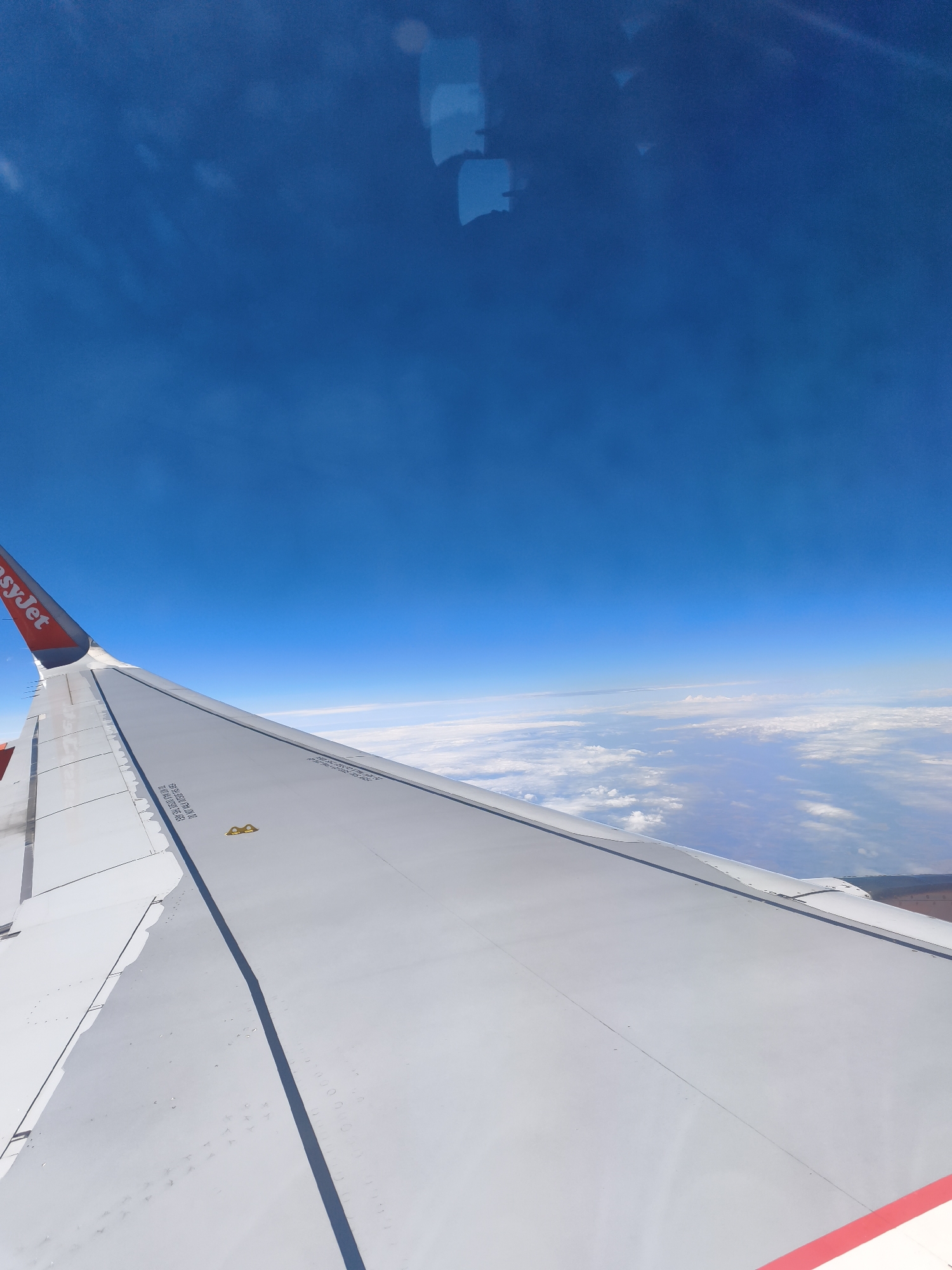 The image shows a view from an airplane window, looking out at the wing and the sky above a cloud layer. The wing is white with red accents and the "easyJet" logo. The sky is a clear, bright blue. The clouds below are white and puffy. The ground is visible through breaks in the clouds, appearing as a patchwork of brown and green. The scene is illuminated by daylight. The image was taken from an airplane flying over Trebbin, Germany.