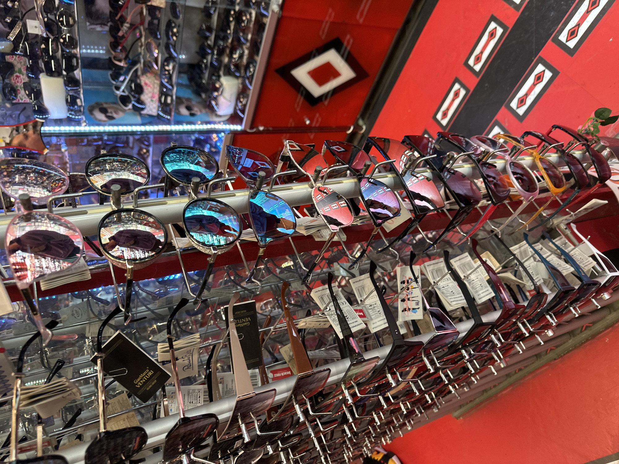 The image shows a display of sunglasses in a store, likely in Pleven, Bulgaria. The sunglasses are arranged on a rack, featuring a variety of styles and colors. The lenses of some sunglasses reflect the surrounding environment, showing the reflections of the store and the outside area. The rack is full and displays price tags.
The background features red shelves with black and white decor. The other shelves in the back contain more sunglass display.