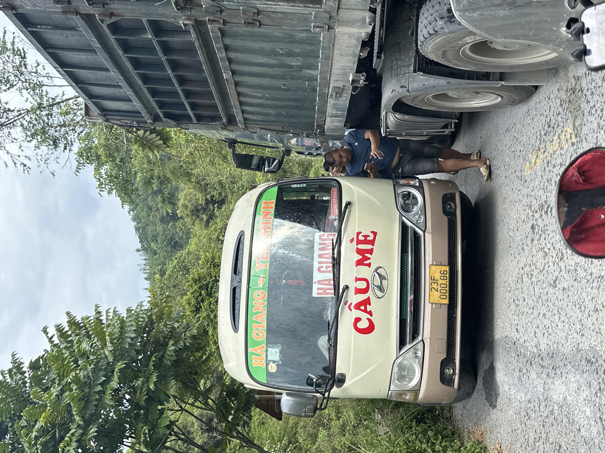 A partially obstructed, medium-shot scene on a road in Bạch Đích Commune, Vietnam, depicts a beige Hyundai passenger van with the inscription "HÀ GIANG - YÊN MINH" and "CẦU MÈ" in red letters on its front windshield, partially obstructed by a large green truck. A man in a blue shirt and grey shorts, wearing sandals, leans towards the front of the van, appearing to inspect the area under the truck's wheels. The truck's tires are visible, indicating its proximity to the van. The van's license plate is visible, reading "23F 000.86". The road surface is asphalt. Green foliage and trees are visible in the background, suggesting a rural or mountainous location. A partial view of a red motorcycle mirror is visible in the right side of the frame.