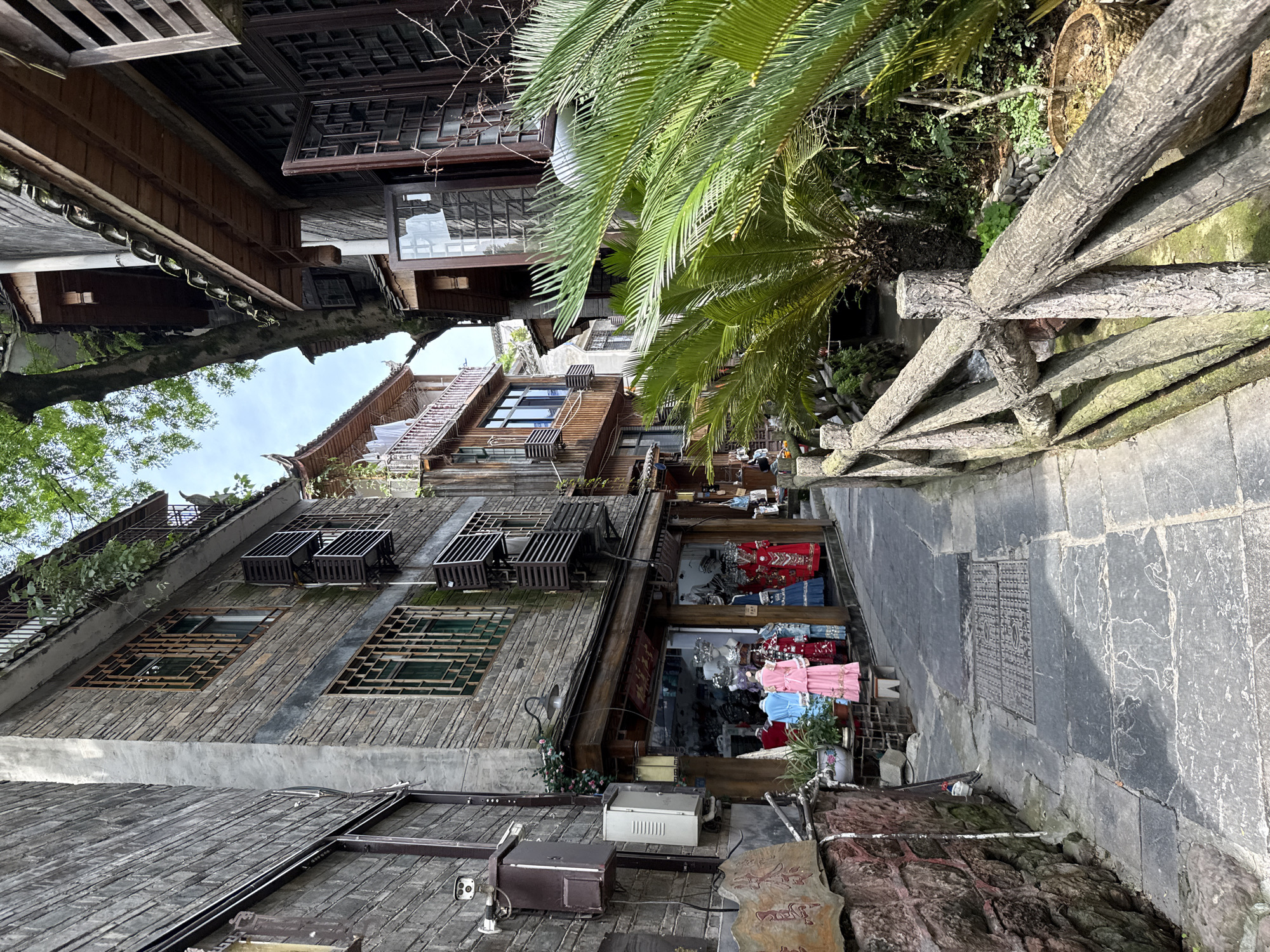 The image depicts a narrow street in Fenghuang, China, lined with traditional wooden buildings. The street is paved with large stone slabs and bordered by a rustic wooden fence on one side. A small shop is visible, displaying clothing items in its window. The buildings are multi-storied with intricate wooden window frames and air conditioning units attached to the exterior walls. Lush greenery, including palm-like plants, overhangs the street, adding a touch of nature to the urban setting. The overall scene captures the unique blend of historical architecture and modern life in Fenghuang.
