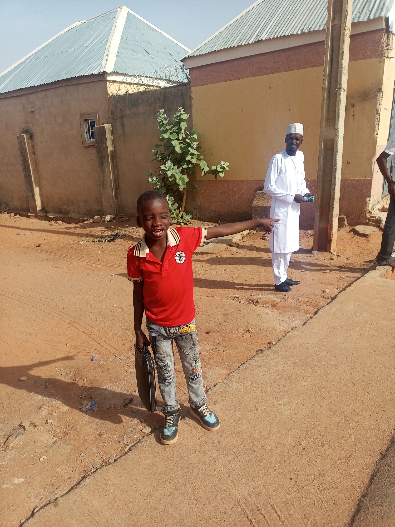 A young boy stands on a dusty, reddish-brown surface in an outdoor setting. He is wearing a red collared shirt, denim jeans with cartoon patches, and sneakers. He holds a black bag in his left hand and points to the right with his right hand. Behind him is an older man wearing a white robe and cap. To the left and behind the boy is a light brown building with a corrugated metal roof and barbed wire along the top of the wall. To the right and behind the older man is a building with a similar roof and a large cement post. There's also a partial figure of a man on the right side of the frame. It is daytime, and shadows are visible. The location appears to be a neighborhood in Tempure, Nigeria.