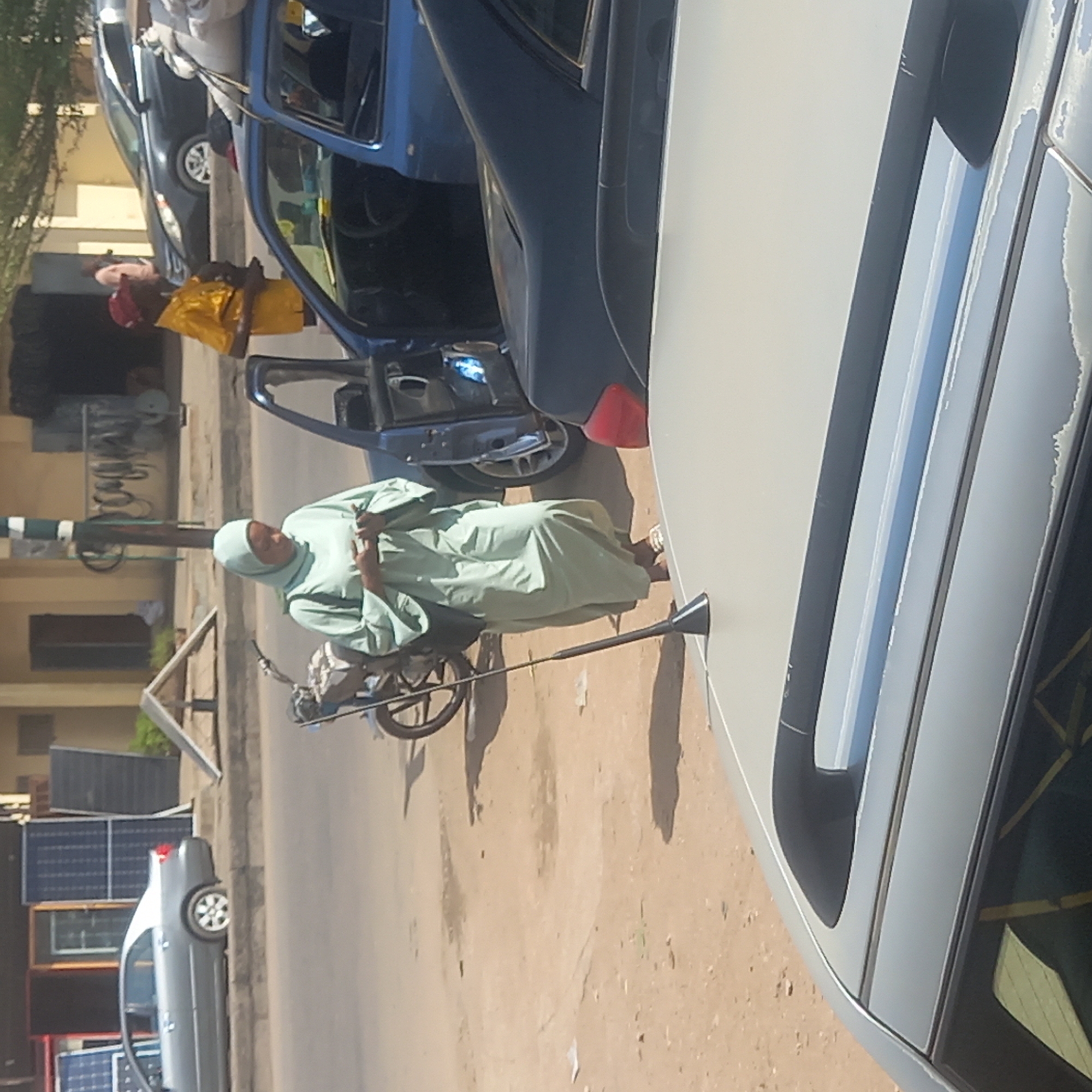The image captures an outdoor street scene in Dadi Riba, Nigeria, under bright daylight. The ground is a light brown, dusty dirt road.
In the mid-ground and slightly to the left, a person wearing a light green traditional long dress (jalabiya or similar) and a matching light green head covering (hijab) stands beside what appears to be a bicycle or motorbike. They are looking towards the viewer, holding a dark-colored smartphone in their right hand, and making a peace sign (V-sign) with the fingers of their left hand.
Further back and to the left, another person is visible, dressed in a yellow top and a red head covering. They are standing near the open front passenger door of a blue sedan car. Behind this blue car, a dark gray or black sedan is parked.
On the far left side of the image, another light gray sedan is parked, and behind it, several dark solar panels are stacked against a building or wall. A tall, dark pole, possibly a street light or utility pole, stands near the person in green.
Part of a light gray car is visible in the extreme foreground on the right, with its roof and a side mirror in view. The scene is illuminated by strong sunlight, creating distinct shadows on the dusty road, indicating a clear, sunny day. No discernible text is visible in the image.