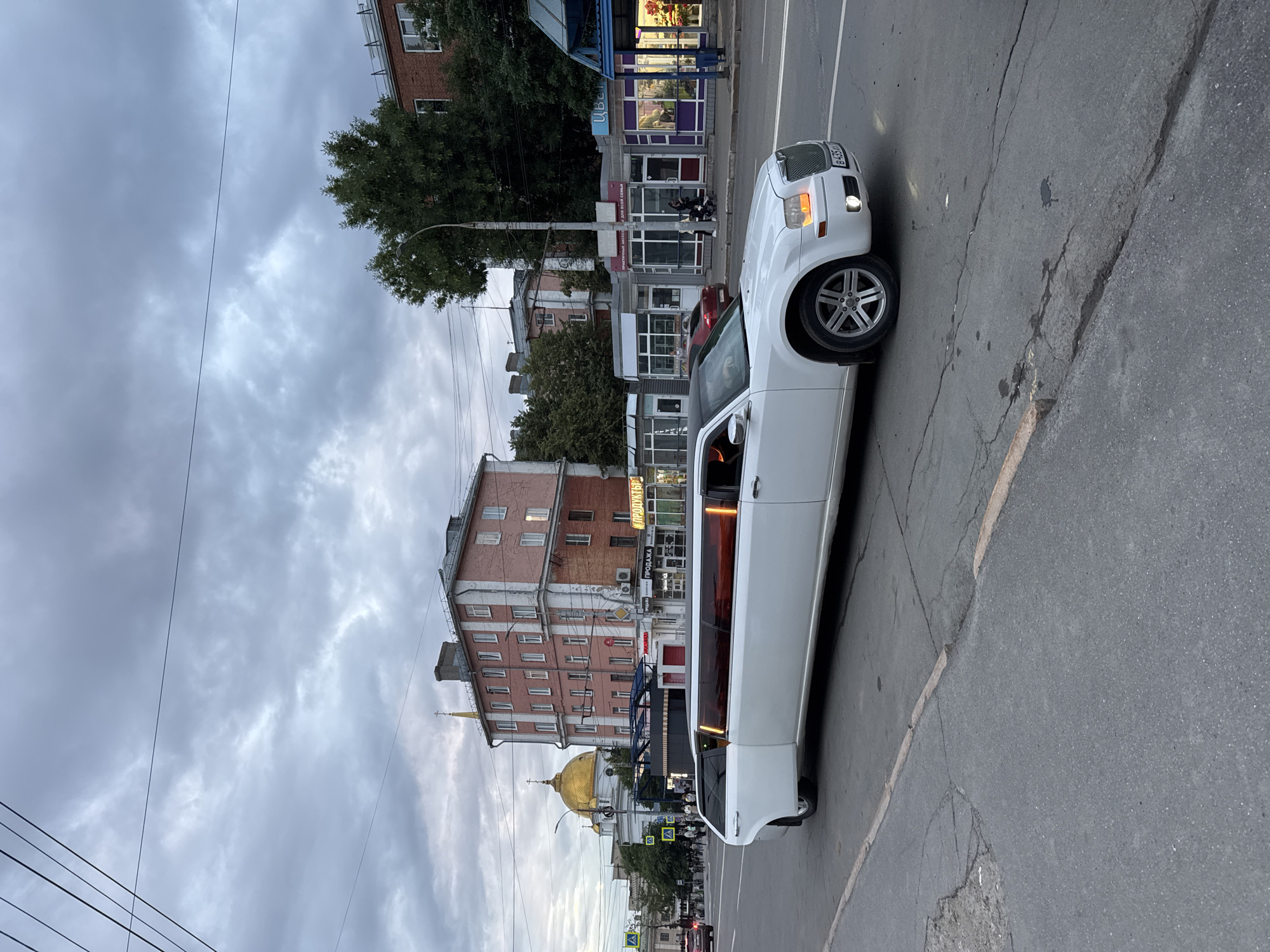 A white limousine drives down a street in Izhevsk, Russia. The sky is overcast and the street is paved with asphalt. Buildings line the street, some of which are shops. A golden-domed church can be seen in the background. There are no people clearly visible in the image.