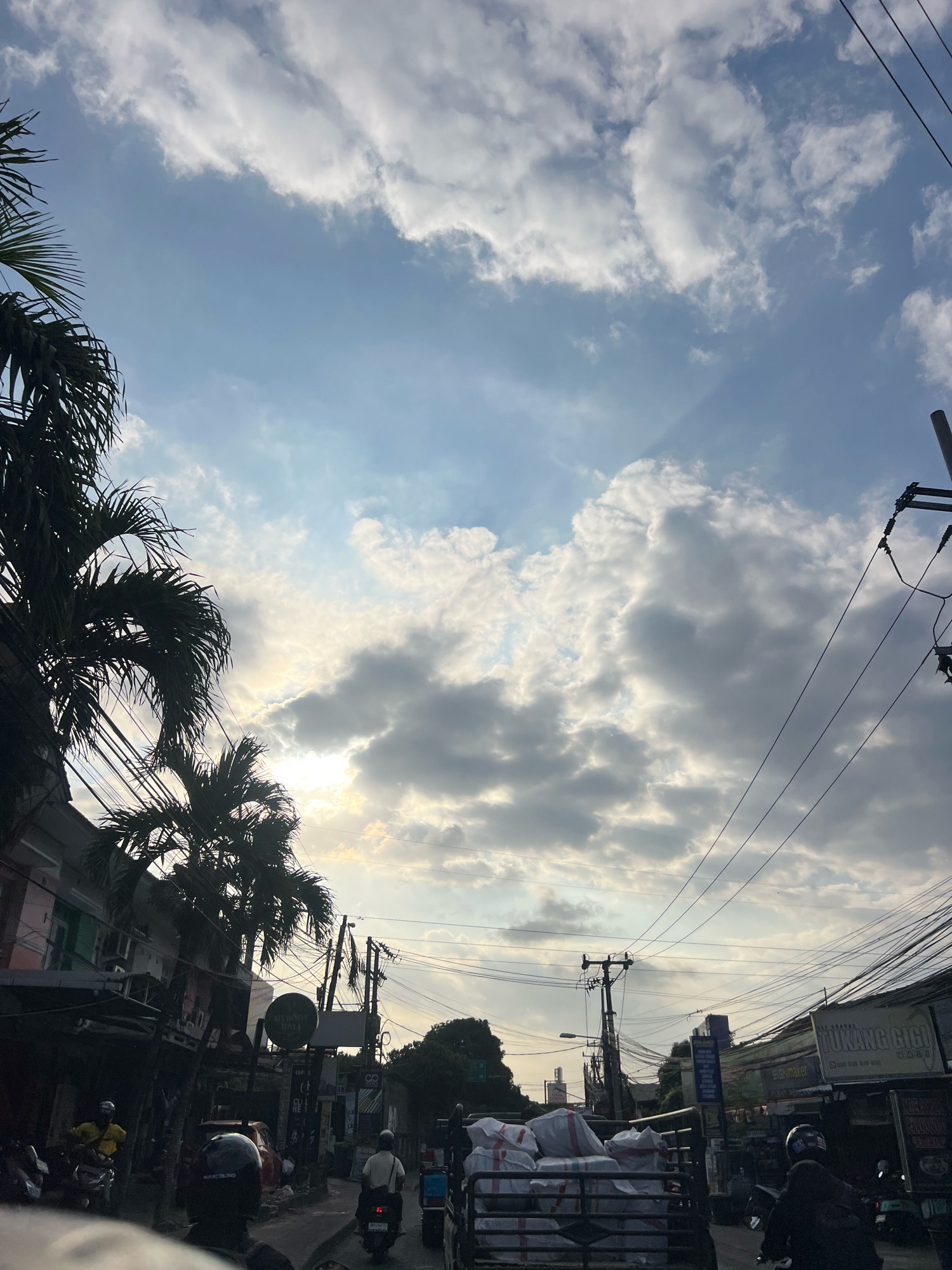 The image depicts a street scene in Denpasar, Indonesia, likely during the late afternoon or early evening. The sky is a mix of blue and grey clouds, with sunlight filtering through, creating dramatic light rays. Palm trees are visible on the left side of the frame, silhouetted against the sky, along with buildings and power lines. The street itself appears to be a busy thoroughfare with several motorbikes and a truck loaded with sacks. People are seen riding motorbikes, some wearing helmets. The overall atmosphere is that of a typical urban environment in Southeast Asia, with a mix of commercial establishments and residential buildings lining the street. The signs visible in the image are in Indonesian, further confirming the location.