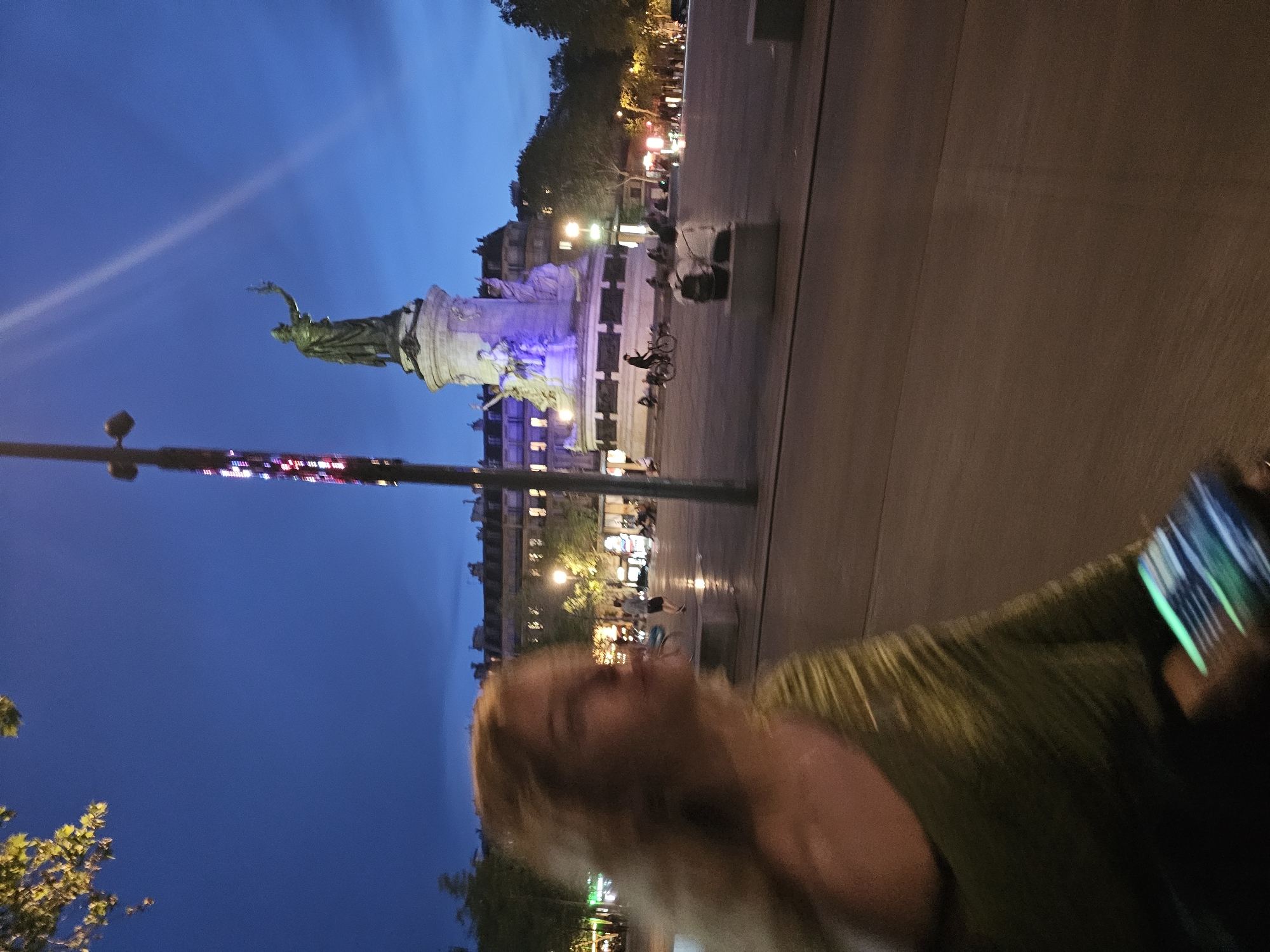 The image captures a nighttime scene in Paris, France, specifically featuring the Place de la République. The statue of Marianne stands prominently in the background, illuminated by a soft purple light. The square is active, with people visible near the monument and along the street. The foreground shows a blurry figure holding a phone, possibly taking a picture. A lamp post with colorful lights is seen to the left. The sky transitions from light blue to a deep dark blue.