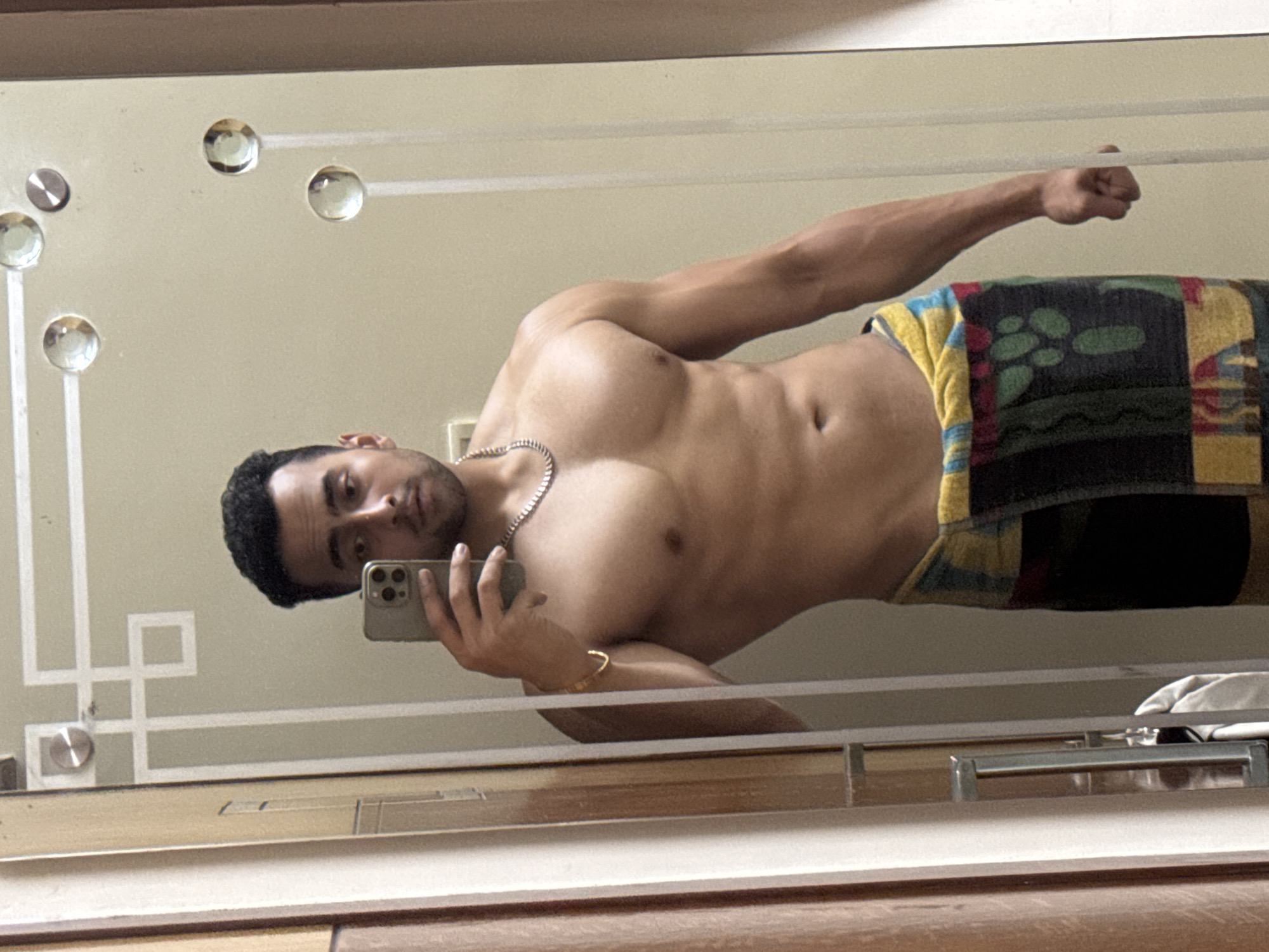 A shirtless man with dark hair and a stubble, wearing a silver chain and a gold bracelet, is holding a smartphone to take a selfie in a mirror. He is partially covered by a colorful towel. The mirror has decorative features, including what appears to be glass or crystal knobs. The background shows what seems to be a wooden surface or furniture. There is no information available in the image to confirm or deny its location in Kolkata, India.