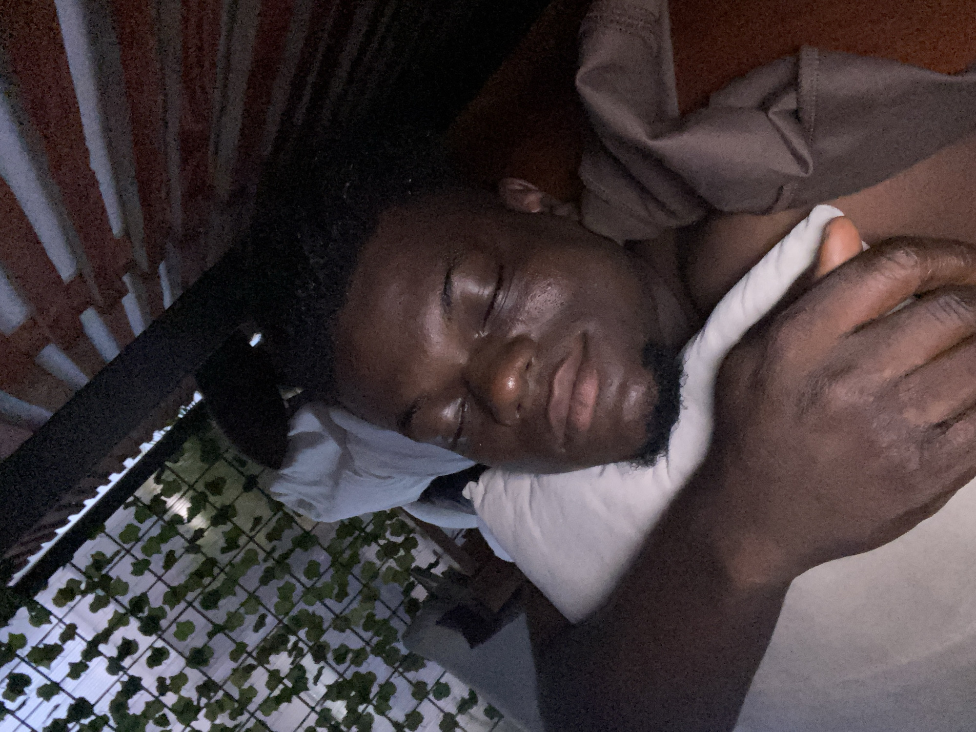 A man with dark skin and short, dark hair is lying on his side on a white pillow. He is facing the camera and his eyes are closed. He has a short beard. His left hand is near his face, resting on the pillow. He is covered by a tan or brown cloth. In the background, there is a reddish-brown and white striped wall on the left, and a wire fence with green ivy. The location is Unknown, Unknown.