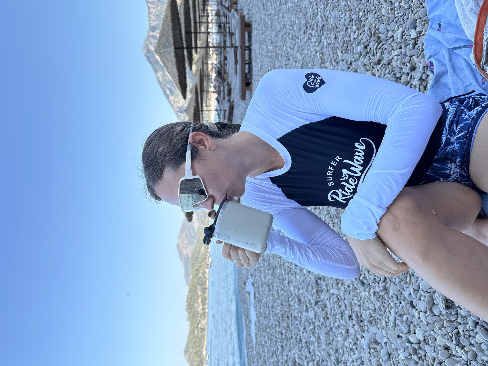 In Fethiye, Turkey, a person wearing a white and black rash guard and dark patterned shorts sits on a pebbled beach. They are holding a light-colored insulated mug to their lips and drinking from it. The person is also wearing large, white-rimmed sunglasses with mirrored lenses. In the background, there are rows of empty lounge chairs and thatched umbrellas, suggesting a beach resort area. The sea is calm and blue, with a mountainous coastline visible in the distance under a clear sky.