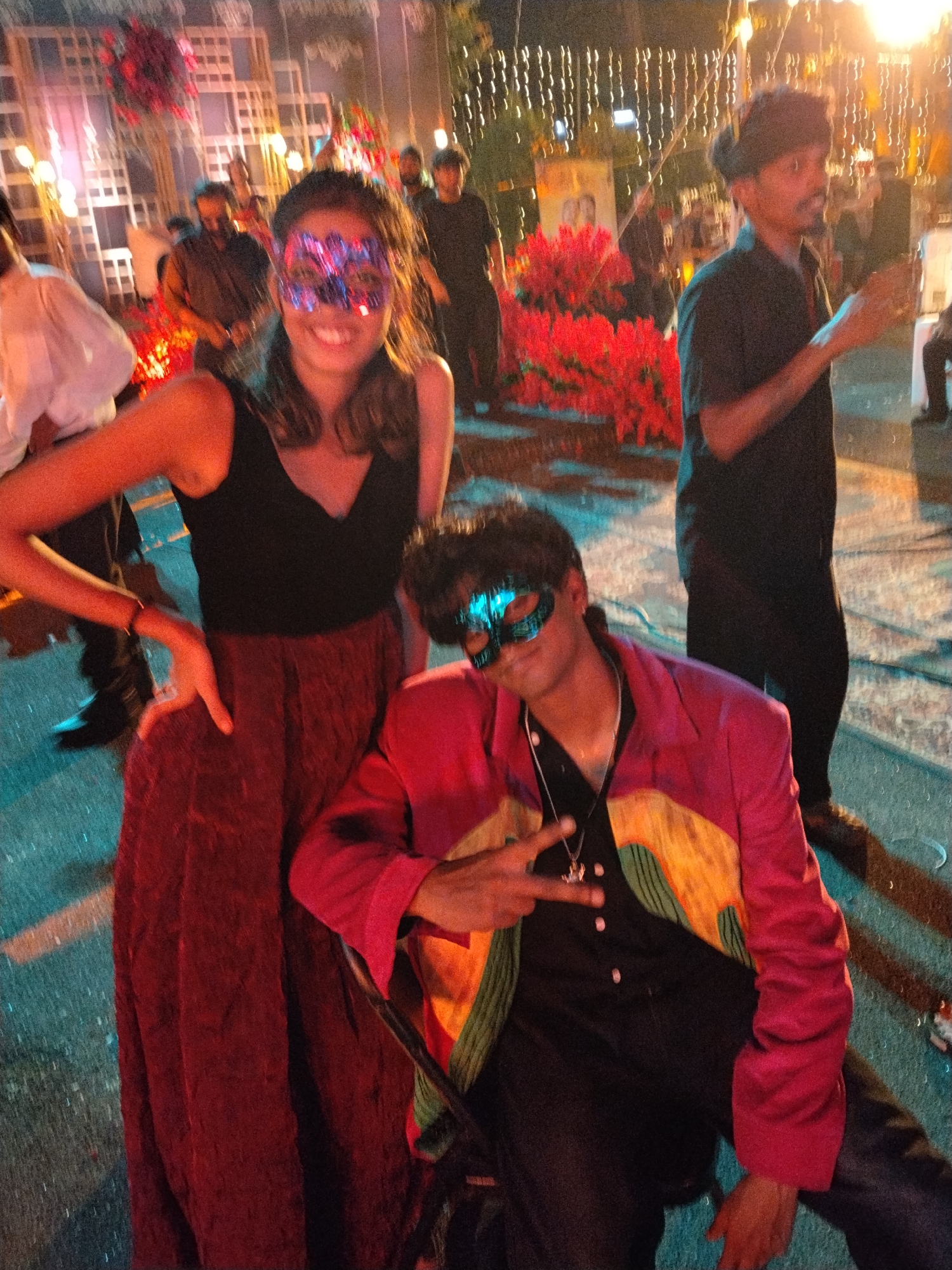 A vibrant nighttime image captures two individuals wearing elaborate masks at an outdoor event in Kalamassery, India. In the foreground, a woman stands smiling, dressed in a black sleeveless top and a long maroon skirt. She wears a striking, multi-colored (blue, purple, red) decorative mask over her eyes, and her left hand is placed on her hip. Beside her, a man is seated or crouched, wearing a black shirt, dark pants, and a distinctive pink, yellow, and green patterned jacket. He sports a blue and green eye mask and makes a peace sign with his right hand, revealing a pendant necklace.
The background is softly blurred but reveals a festive atmosphere. Numerous warm-toned string lights hang overhead, creating a bokeh effect, and bright red artificial floral or bush decorations are prominently featured. A grid-like illuminated structure is visible further back. Several other blurred figures are present, indicating a social gathering or party. The ground appears wet and reflects the ambient light, particularly with blue and teal hues.
The overall mood is joyous and celebratory, emphasized by the subjects' poses and the vibrant decorations. The scene is illuminated entirely by artificial light, clearly indicating it's nighttime. No discernible text is visible in the image.