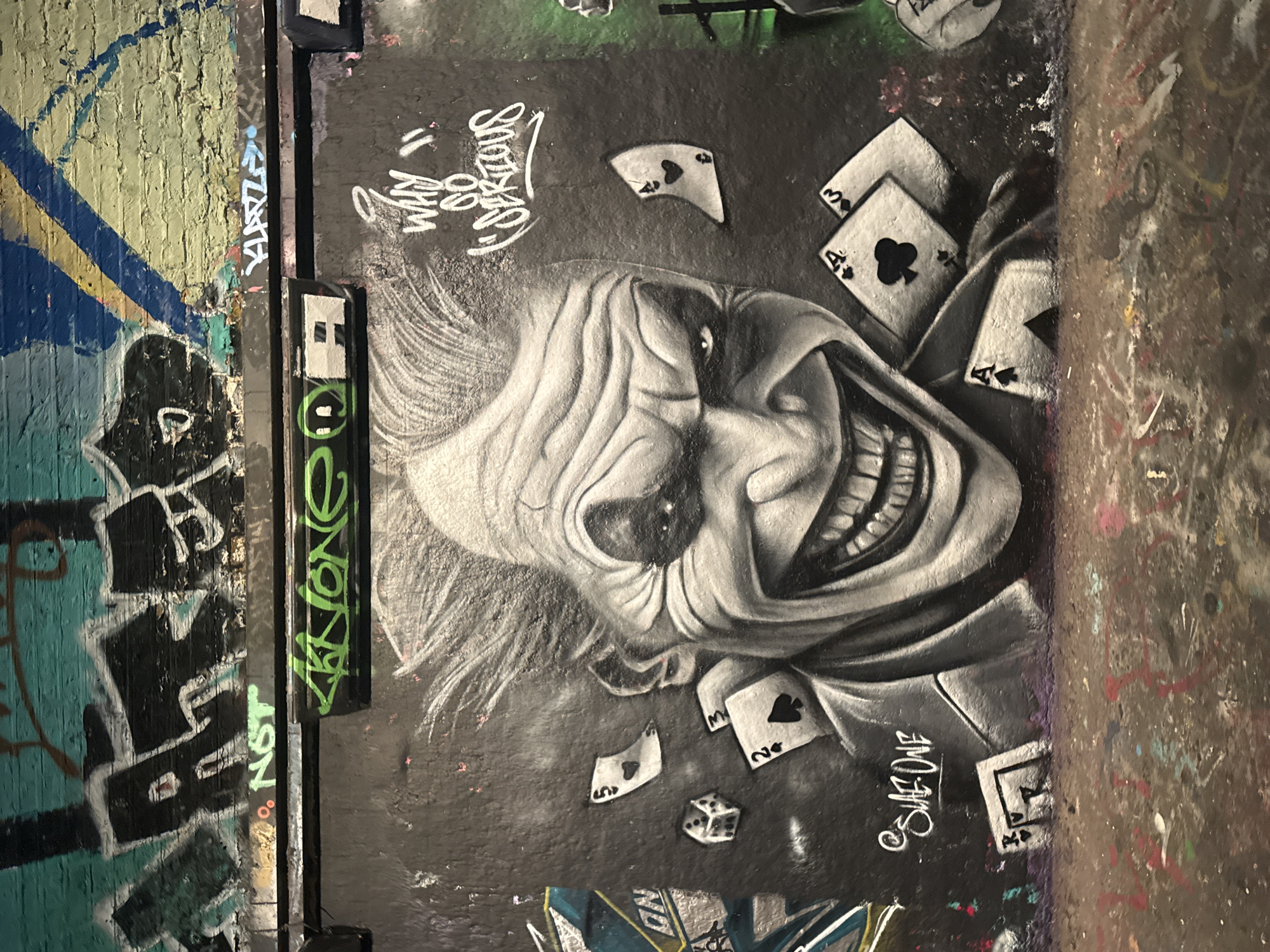 The image shows graffiti art on a wall, likely located in an urban environment. The main subject of the graffiti is a black and white depiction of the Joker character from the Batman series, complete with a wide, exaggerated grin. Playing cards are scattered around the Joker's face, creating a chaotic atmosphere. Other graffiti tags and abstract designs are present on the surrounding wall, adding to the urban aesthetic. It is not possible to discern the exact location within London.