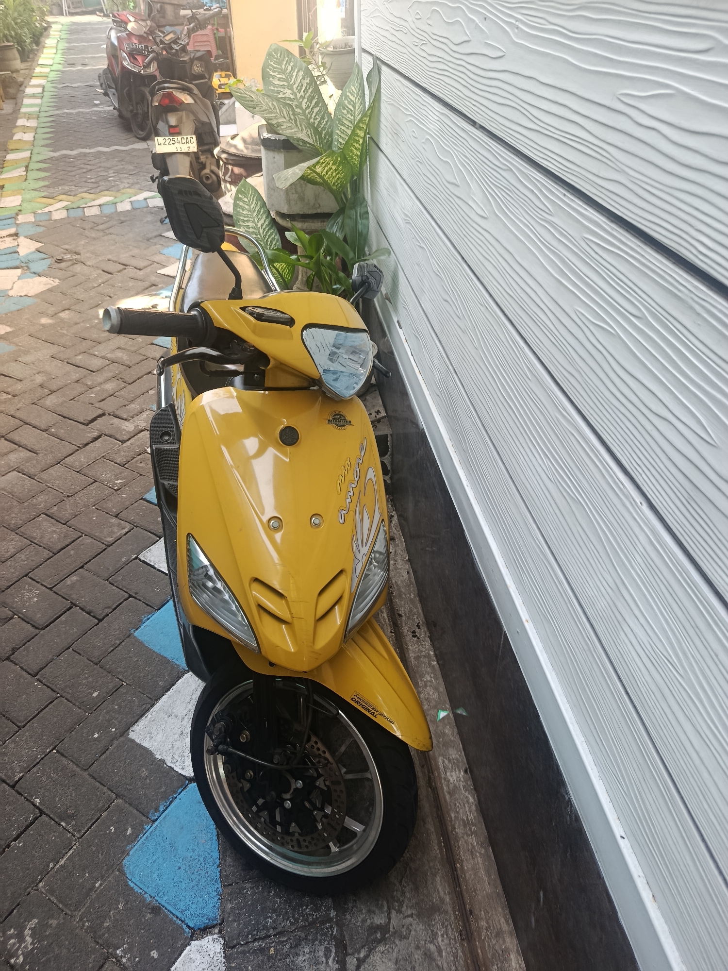 A yellow Yamaha Mio scooter is parked on a brick-patterned walkway with blue and white painted markings. The scooter is positioned diagonally, facing towards the viewer's left, with its front wheel prominently in the foreground. To the right of the scooter is a light-colored, horizontally paneled wall with a wood-grain texture. Behind the scooter, in the background, are other motorcycles parked in a narrow alleyway. The alleyway's walkway is illuminated by sunlight, creating shadows. This scene is located in Surabaya, Indonesia.