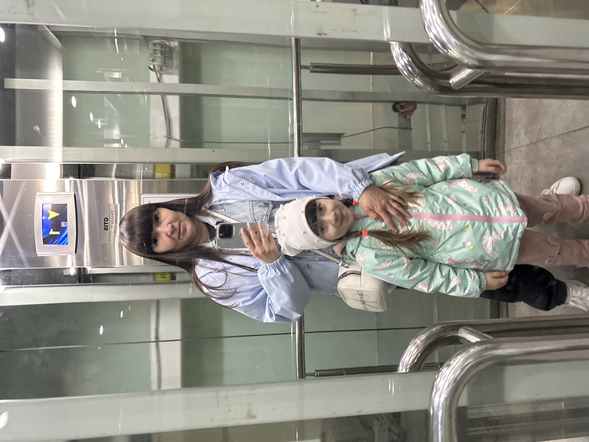 The image shows a woman and a young girl standing in an elevator. The woman is holding a smartphone and appears to be taking a selfie. The elevator's control panel is visible, indicating they are on the first floor. The location is potentially in Bolshoye Savino, Russia, but this can't be determined from the image. The elevator has glass walls. The woman is wearing a light blue jacket. The girl is wearing a white hat and a light green coat with a unicorn pattern. She is also wearing pink pants. The elevator has metal handrails.