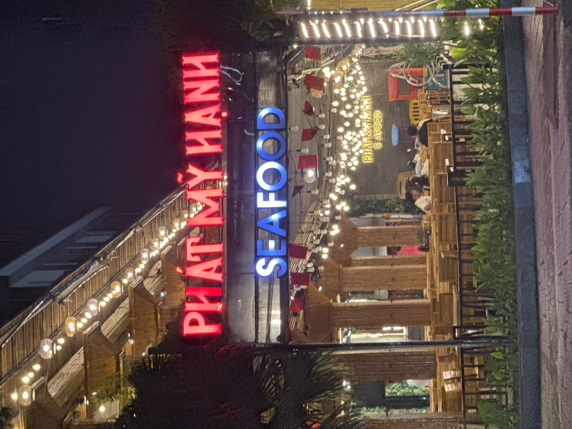 The image captures a nighttime scene in Đà Nẵng, Vietnam, featuring the illuminated signage of a seafood restaurant named "PHÁT MỸ HẠNH SEAFOOD". The red and blue neon lights of the sign are prominently displayed. The restaurant appears to be an open-air establishment with multiple levels and bamboo-like structures, adorned with decorative lights. Diners are visible at some tables. The surrounding environment includes lush greenery and trees, contributing to the ambiance. A section of a pathway or sidewalk is seen on the right edge of the image.