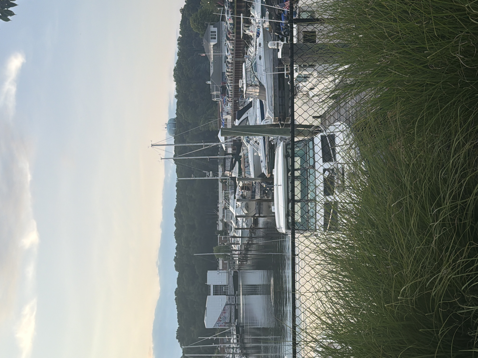 The image depicts a harbor in the City of Glen Cove, United States of America. Several boats, primarily white motor yachts and sailboats, are docked in the water. The foreground is dominated by tall, green grass and a chain-link fence. In the background, a modern white building with large windows is visible on the left, reflecting in the water. Further back, a dense line of trees forms a backdrop, with a light blue water tower perched on top of a hill. The sky is partly cloudy with a soft, diffused light, suggesting it is either early morning or late afternoon. The overall scene conveys a sense of tranquility and a typical day at a marina.