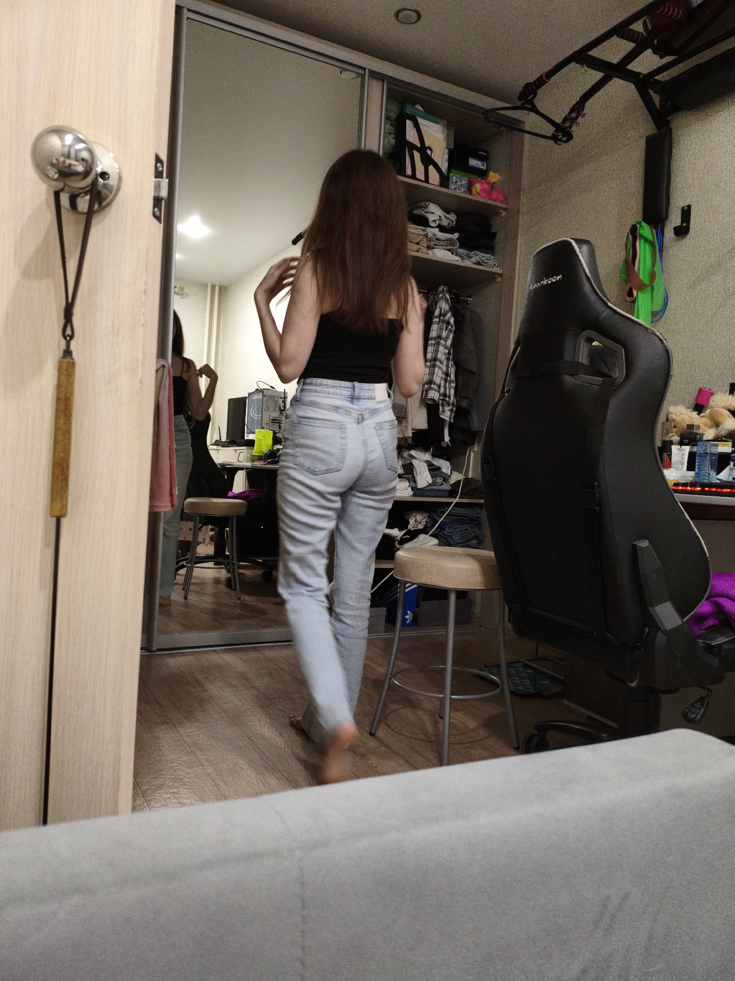 The image depicts an indoor scene, likely a bedroom or living space in an apartment. A young woman with long brown hair is standing in the foreground, facing away from the camera and towards a full-length mirror. She is wearing a black sleeveless top and light blue denim jeans. Her right hand is raised, touching her hair. The mirror reflects the woman and the room behind her.
The room appears to be somewhat cluttered. To the left of the mirror is a wooden wardrobe or closet door with a silver handle and a hanging decorative item. To the right of the woman, the mirror reflects a closet area with shelves containing clothing and various items. Adjacent to the closet is a black gaming chair. Further to the right, a desk is visible with a computer monitor, a yellow object, and other miscellaneous items. A stool is positioned in front of the desk. The floor is made of wooden planks. The overall lighting suggests it is daytime, possibly artificial light from the room. There are no explicit indicators of the specific location within Chelyabinsk, Russia, beyond the context of a domestic interior.