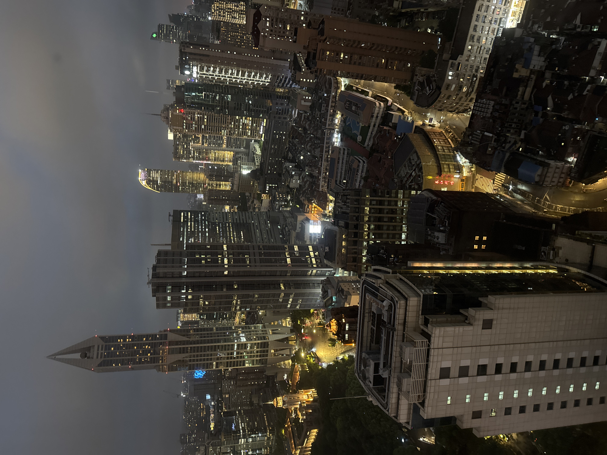This is a high-angle, nighttime aerial photograph of a dense urban cityscape in Shanghai, China. The image captures a panorama of illuminated skyscrapers, buildings, and city lights under a cloudy sky.
In the foreground, a prominent, light-colored skyscraper with a patterned facade is partially visible, showing rows of lit windows. To its right, a darker, glass-faced building with vertical yellow lighting stands tall.
Further into the cityscape, a very tall, distinct skyscraper with a pointed, almost spire-like top, illuminated by internal lights and some red accent lights, dominates the left side of the frame. To its right are numerous other high-rise buildings of varying heights and architectural styles, many displaying illuminated windows.
In the mid-ground, a more historic-looking building complex with a clock tower and warm yellow lighting is visible, situated near a street with passing vehicles and streetlights. Surrounding this are various other buildings, some with curved facades and illuminated signage, indicating commercial activity. The lower portion of the image reveals a cluster of lower-rise buildings with dark roofs, densely packed together, suggesting older urban development.
The overall impression is of a sprawling, vibrant metropolis at night, characterized by a mix of modern high-rise architecture and older structures, all aglow with artificial light.