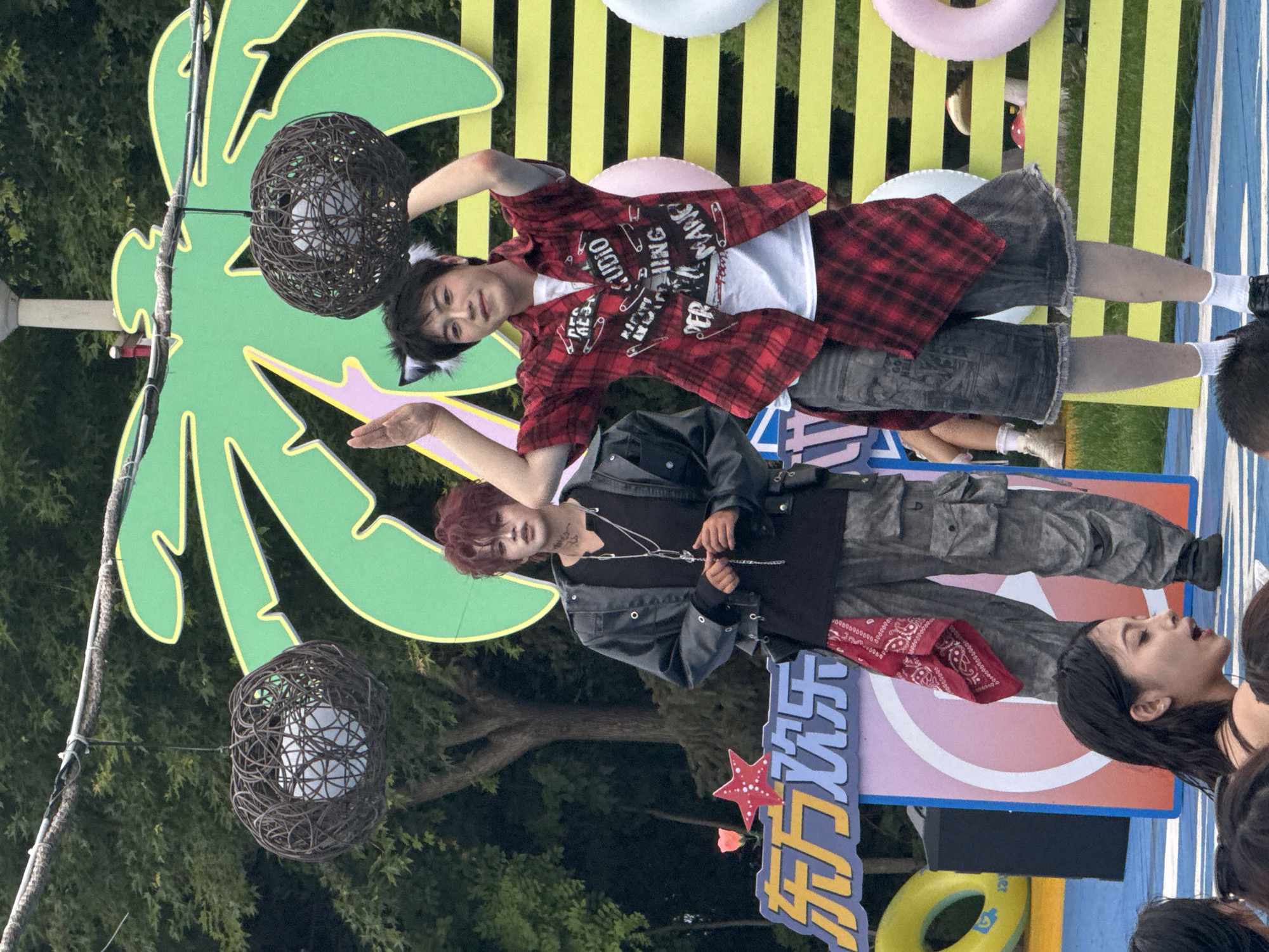 Two young people are standing on a platform decorated with a palm tree motif and yellow and green stripes. The person on the left has reddish-brown hair and is wearing a black leather jacket over a black t-shirt, grey cargo pants, and a red bandana tied around their waist. The person on the right has black hair and is wearing a red plaid shirt with patches and a white t-shirt underneath, grey denim shorts, and white socks. They are both wearing black shoes. The person on the right is also wearing cat-ear headwear. In the foreground, out of focus, is the head and shoulders of a young woman. Behind the platform, a sign with Chinese characters is visible. In the background, there are trees and an overcast sky. The scene appears to be outdoors, possibly at an event or festival. There is no information in the image to suggest the location is in Taihe District, China.