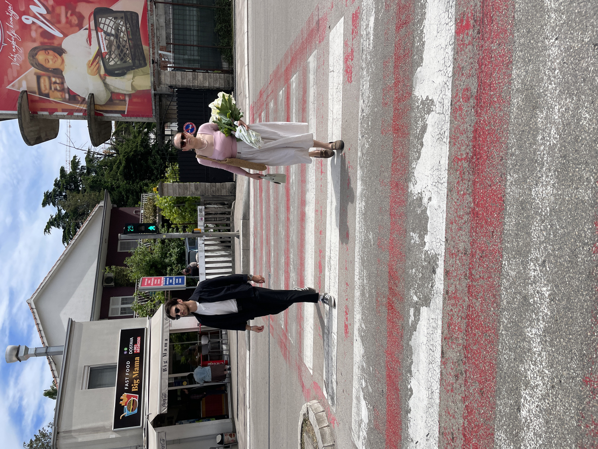 Two individuals are crossing a zebra-striped crosswalk. The person at the top of the image, a woman, is wearing a light pink top, a pearl necklace, a light-colored skirt, sunglasses, and carrying a bouquet of white flowers. She also has a woven straw bag and a small white object in her other hand. The person at the bottom of the image, a man, is wearing a black suit jacket and pants, a white t-shirt, sunglasses, and black and white sneakers.
The crosswalk is painted with alternating white and red stripes. The street appears to be gray asphalt.
Behind the man is a building with a sign that reads "FAST FOOD" and "Big Mama" along with a telephone number. The sign also features an image of French fries in a container. To the left of the sign is a Coca-Cola branded umbrella.
Behind the woman, there is a building with trees and bushes. A green traffic light is visible. Above the trees, a billboard is visible. It depicts a woman in a white coat holding a shopping basket and text "Vaz najbolji komšija!".
The scene appears to be in Budva, Montenegro, based on the local business name and potential Cyrillic writing on the billboard.