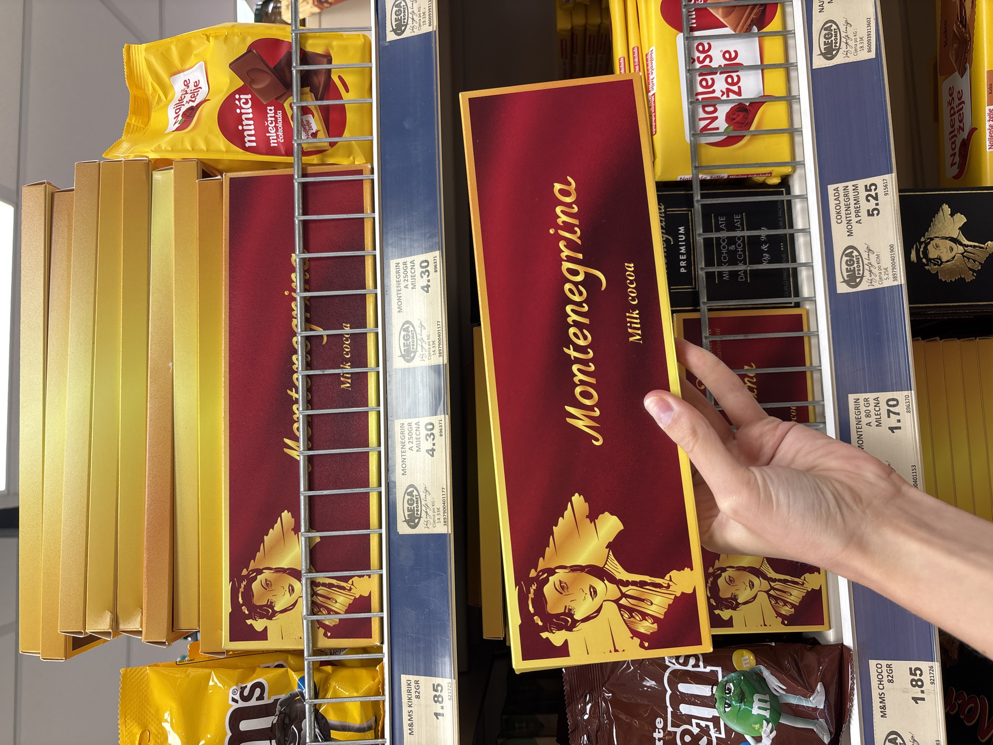 The image shows a shelf in a supermarket, likely in Boreti, Montenegro, stocked with various chocolate products. In the foreground, a person's hand is holding up a rectangular box of "Montenegrina" milk cocoa chocolate. The box is dark red with gold lettering and features an illustration of a woman. Behind this box, several other identical boxes are stacked.
To the left of the Montenegrina boxes, a yellow bag of M&M's is visible. Below the Montenegrina boxes, there are price tags. One tag displays "M&MS KIKIRIKI 82GR" priced at "1.85". Another tag shows "MONTENEGRIN A 250GR MIJECNA" priced at "4.30". Further down the shelf, another price tag indicates "M&MS CHOC 82GR" at "1.85", and a "MONTENEGRIN A 80 GR MLEČNA" at "1.70".
To the right of the Montenegrina boxes, yellow packaging with the text "Najlepše želje" and images of chocolate bars is present. A larger yellow bag with "Najlepše želje" and "minići mlečna cokolada" is also visible. In the bottom right, another "Najlepše želje" product is displayed. The environment appears to be a retail store with fluorescent lighting.