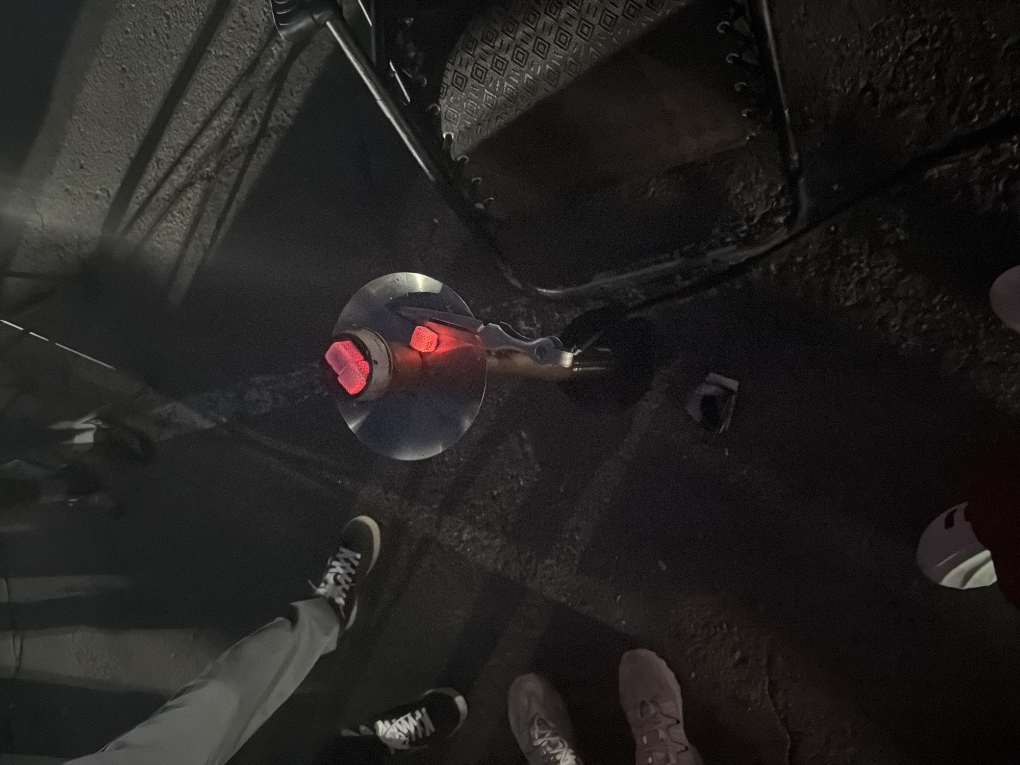 An aerial-view shot depicts a dimly lit outdoor scene, likely a city street or alleyway in Unknown, Unknown. A metal hookah with glowing coals sits on the ground near a folding chair. Three pairs of feet are visible: one wearing grey pants and black-and-white sneakers, one wearing dark sneakers, and another with light-colored sneakers. A glimpse of a shoe is seen near the right edge, and an indiscernible dark object is nearby. The environment appears to be paved.