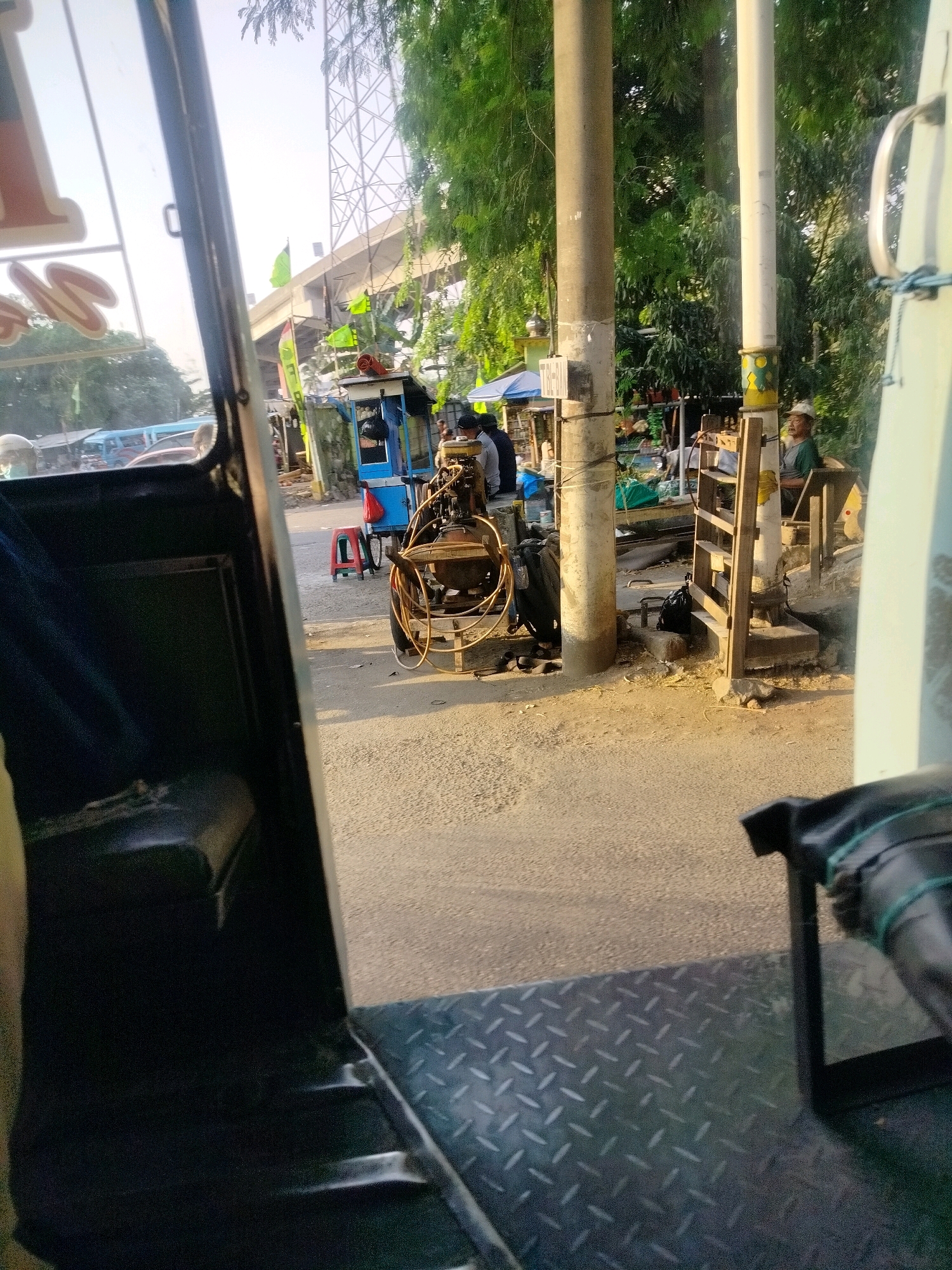 The image captures a street scene in East Jakarta, Indonesia, from the perspective of someone inside a three-wheeled vehicle, possibly a becak motor or a similar local transport. The foreground shows the interior of the vehicle, including a black seat and a diamond-patterned metal floor. The midground and background reveal a street with various stalls and structures.
On the right side of the street, a man wearing a cap and a green shirt is seated in what appears to be a wooden chair at a stall. Beside him is a wooden shelf structure. Two large concrete poles, likely utility poles, stand prominently in the midground. One is a light gray concrete, and the other is painted yellow and green at its base.
To the left of the poles, a blue food cart or stall is visible, with a red plastic stool placed in front of it. A person in dark clothing is standing behind the cart. A small, dark-colored generator with hoses is positioned near the cart and the concrete poles. Green flags are visible on poles behind the stalls, and a large electricity pylon or tower is seen in the distance, beyond an elevated road structure.
The street surface is paved asphalt, with patches of dirt and debris. Trees with dense green foliage line the background, suggesting a relatively tropical environment. Sunlight casts shadows, indicating it's likely late afternoon or early morning. The overall impression is of a bustling roadside area in a developing urban setting.