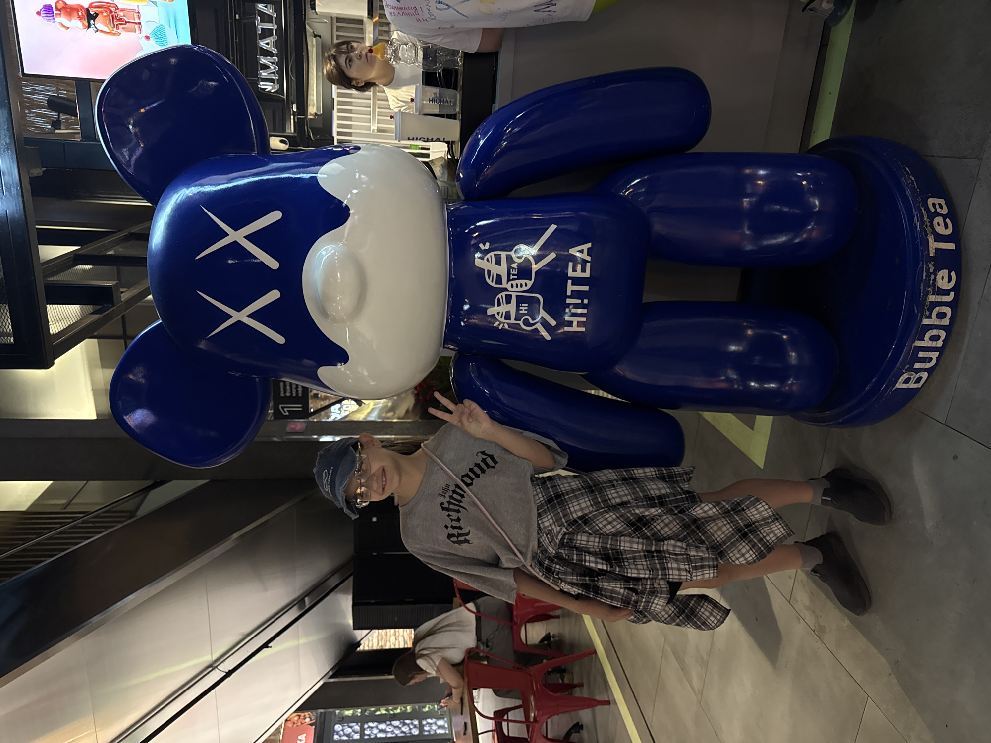 The image depicts a scene in what appears to be a shopping mall. A person with short hair, wearing glasses, a gray t-shirt with "Richmond" printed on it, a plaid skirt, and sneakers, is posing next to a large blue and white bear-shaped sculpture, decorated with the logo "Hi!TEA". The sculpture is placed on a blue base with the words "Bubble Tea" written on it. In the background, there is an escalator, a store or a restaurant with tables and red chairs, and a woman standing behind a counter. Text is displayed on multiple signs. Based on the Cyrillic text that is visible, it is possible the image was taken in Russia, potentially in Moscow.