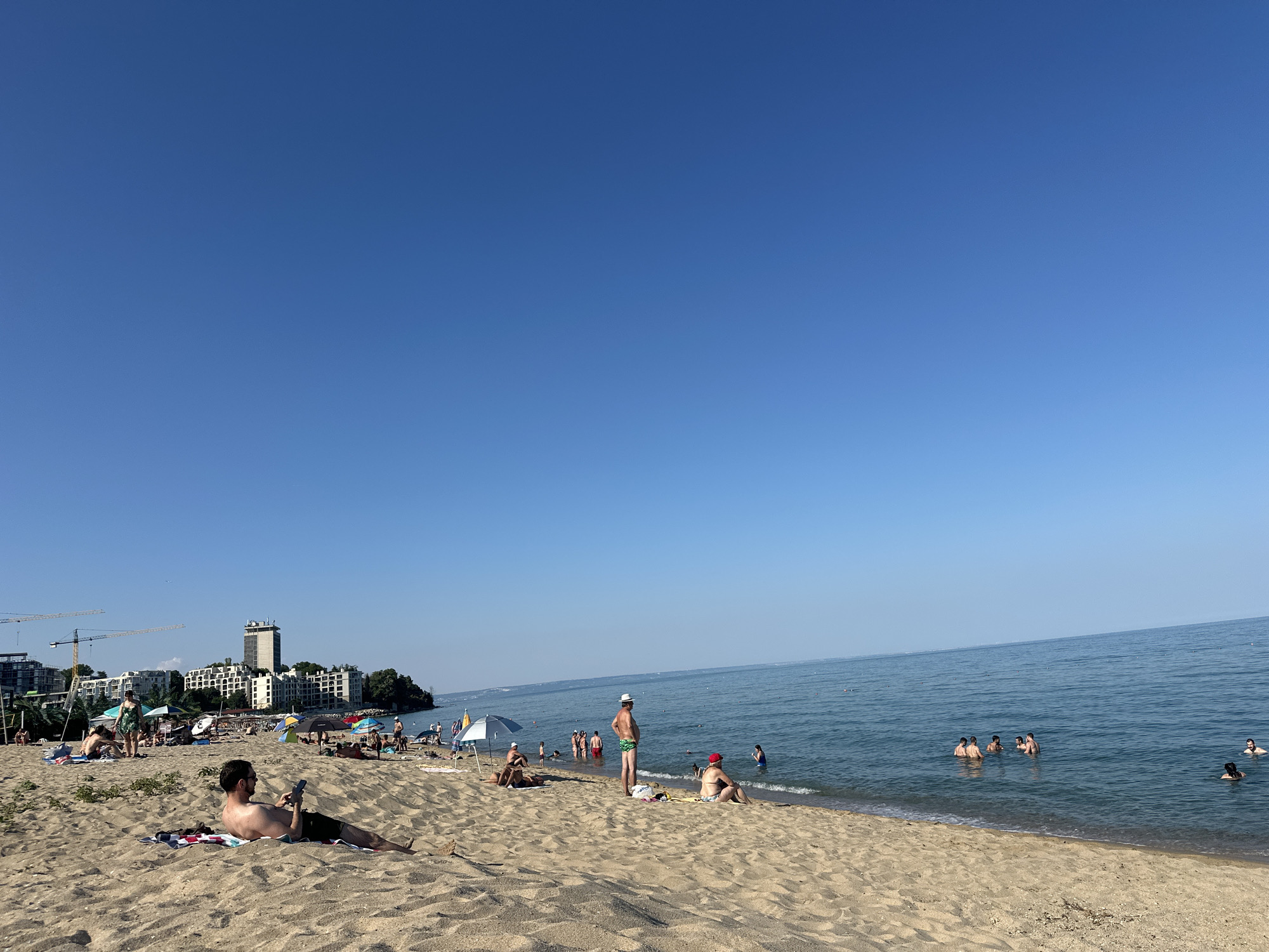 This is a wide shot of a crowded beach on a clear, sunny day. The sky is a vibrant blue with no clouds. The sand is light brown and appears dry. Several people are scattered across the beach, some lounging on towels, others wading or swimming in the calm, blue ocean. In the foreground, a man in sunglasses lies on his back on a towel, looking at his phone. Further back, more people are visible under beach umbrellas.
Along the coastline in the background, there are several buildings, including a tall, modern hotel and other apartment-like structures. Construction cranes are also visible to the left, suggesting ongoing development. The sea stretches to the horizon, meeting the clear blue sky. The overall atmosphere is one of relaxation and a typical summer day at the beach. The time of day appears to be midday or early afternoon, given the bright sunlight and clear sky.