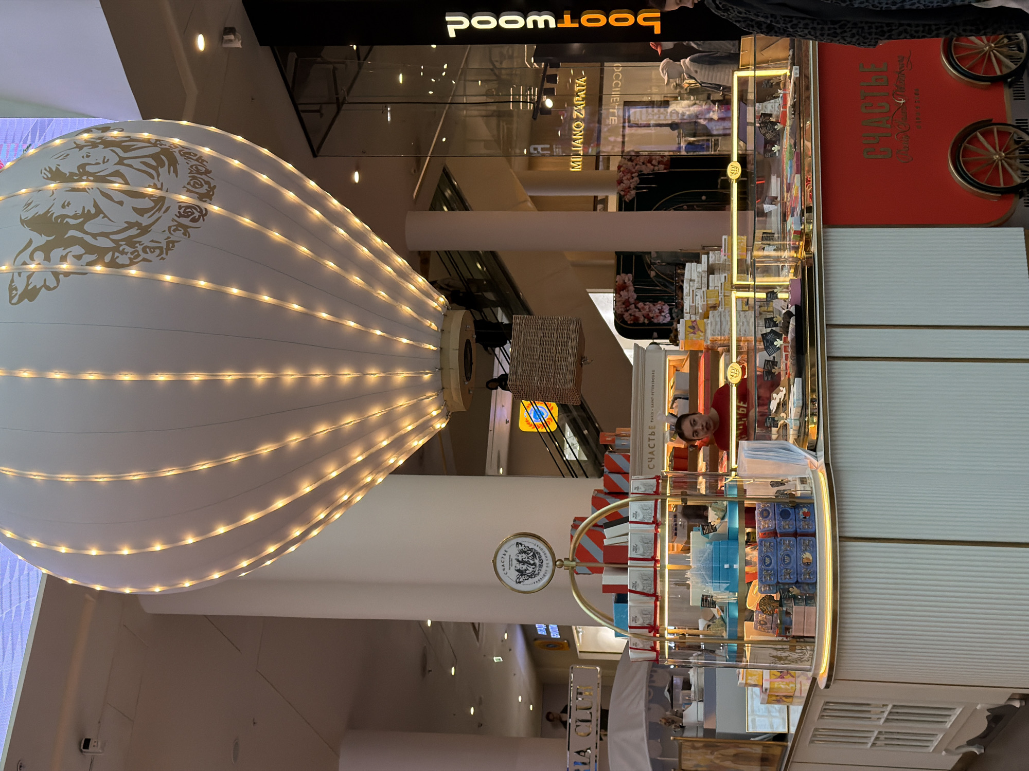 The image depicts an indoor view, seemingly inside a shopping mall in Saint Petersburg, Russia, featuring a large, white, hot air balloon-shaped art installation with gold accents and illuminated with white lights. Below the balloon, there's a kiosk or shop, possibly a retail location for a brand named "СЧАСТЬЕ" (Happiness), displaying various merchandise in glass showcases. A person wearing a red shirt is visible inside the kiosk. In the background, the "Poomwood" sign and other shops are present, and an upper level of the mall is visible, suggesting a multi-story shopping complex.