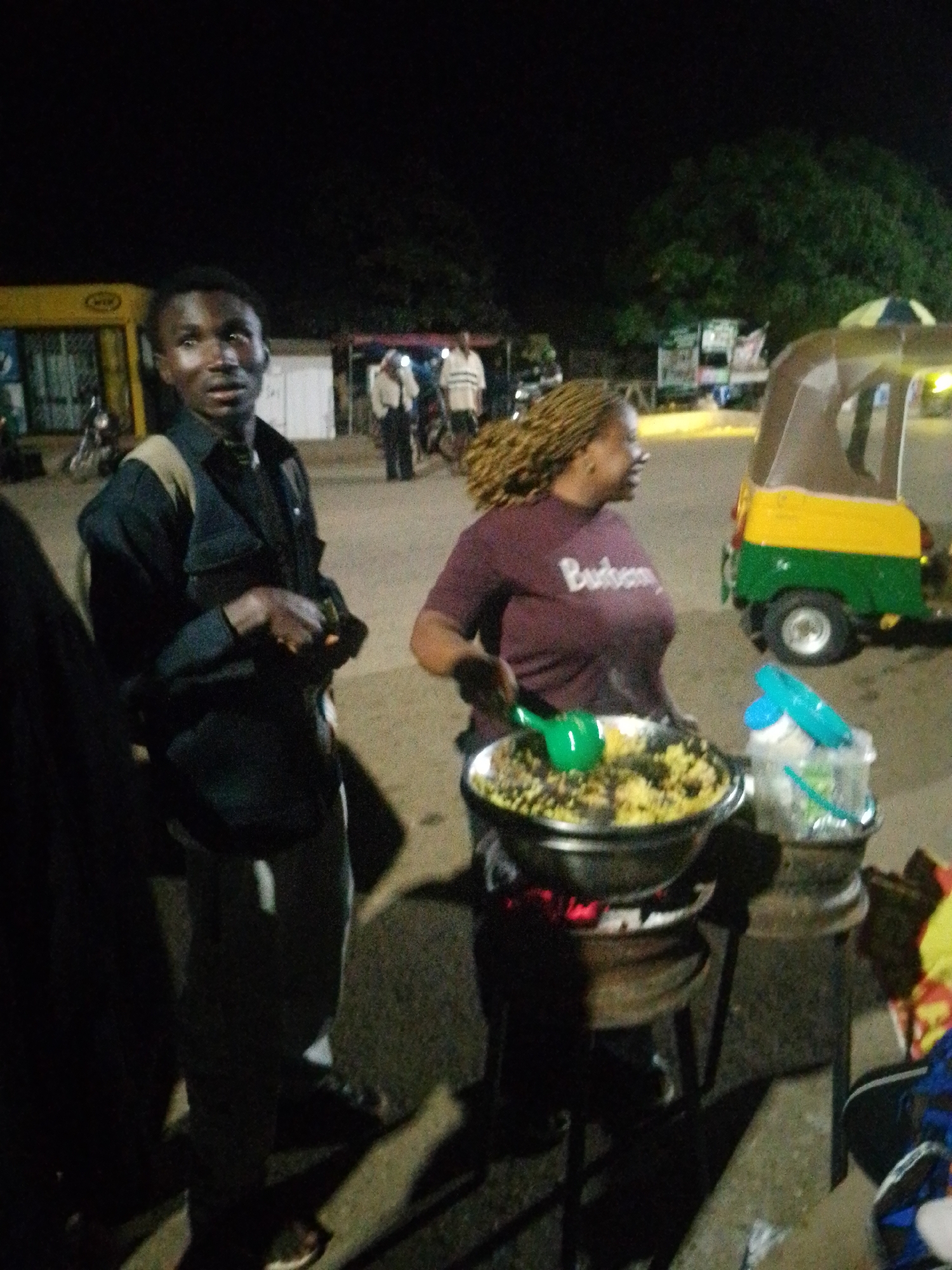 The image depicts an outdoor scene at night. In the foreground, a man wearing a dark jacket and a light-colored undershirt is standing. To his right, a woman with braided hair, wearing a maroon t-shirt with "Burberry" written on it, is serving food from a large metal bowl. The food appears to be a yellow grain dish with other ingredients. The food is being cooked over coals, which are visible beneath the bowl. Next to the food preparation area, there are plastic containers with what appear to be beverages and utensils. In the background, an auto-rickshaw, painted yellow and green, is parked. Several other people are visible in the distance, along with buildings and trees, suggesting an urban environment. The lighting is dim, characteristic of nighttime.