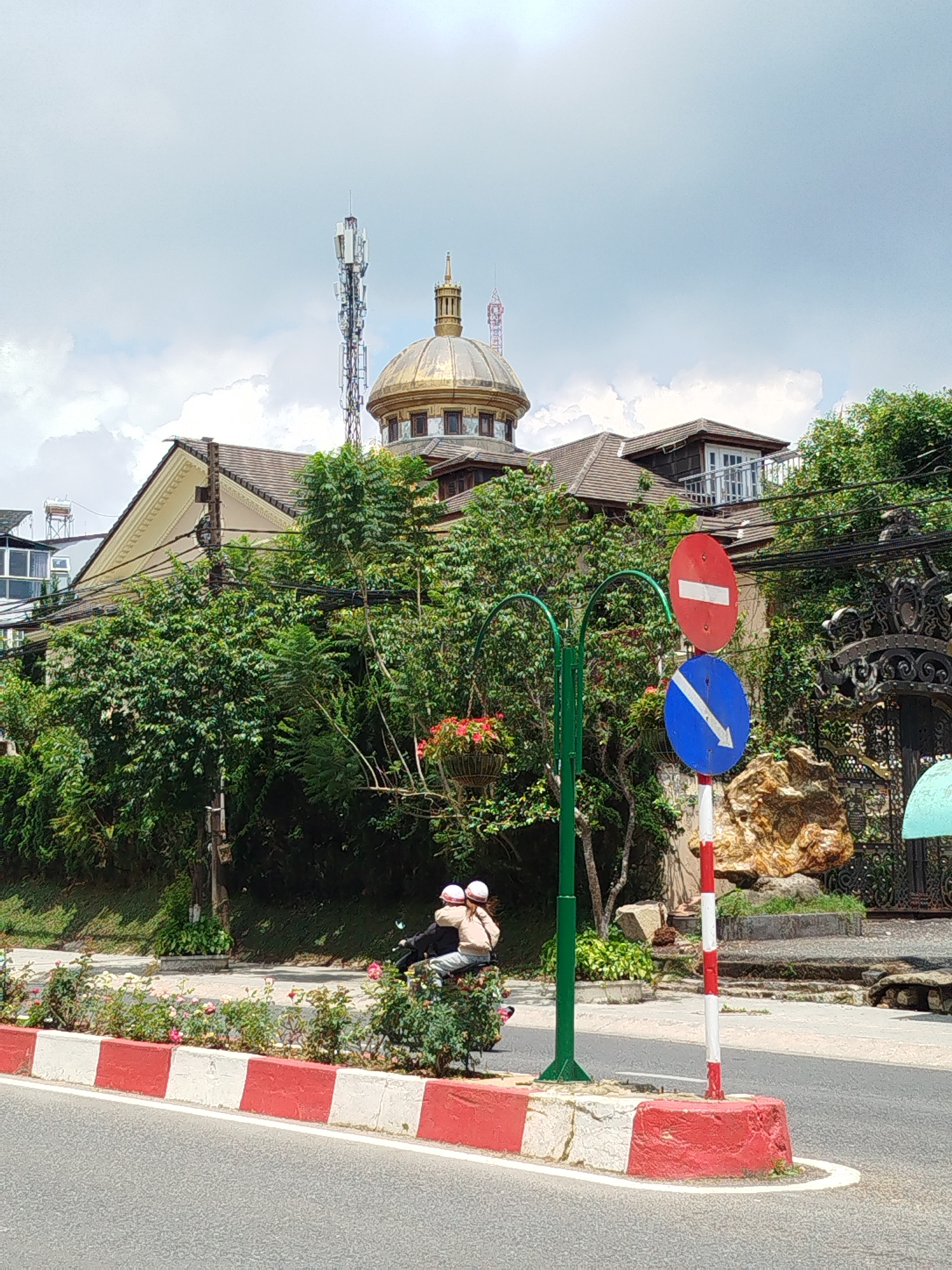 The photograph is taken at street level, likely in Da Lat, Vietnam, and depicts a street scene with a view of a building featuring a gold dome. A red and white curb lines the road, and a signpost with a red "no entry" symbol and a blue arrow points to the left of the frame. Two individuals wearing helmets are riding a motorbike behind a row of roses, obscured by the foliage. The building in the background is surrounded by greenery, with a telecommunications tower visible behind it. The sky is overcast with white and gray clouds.