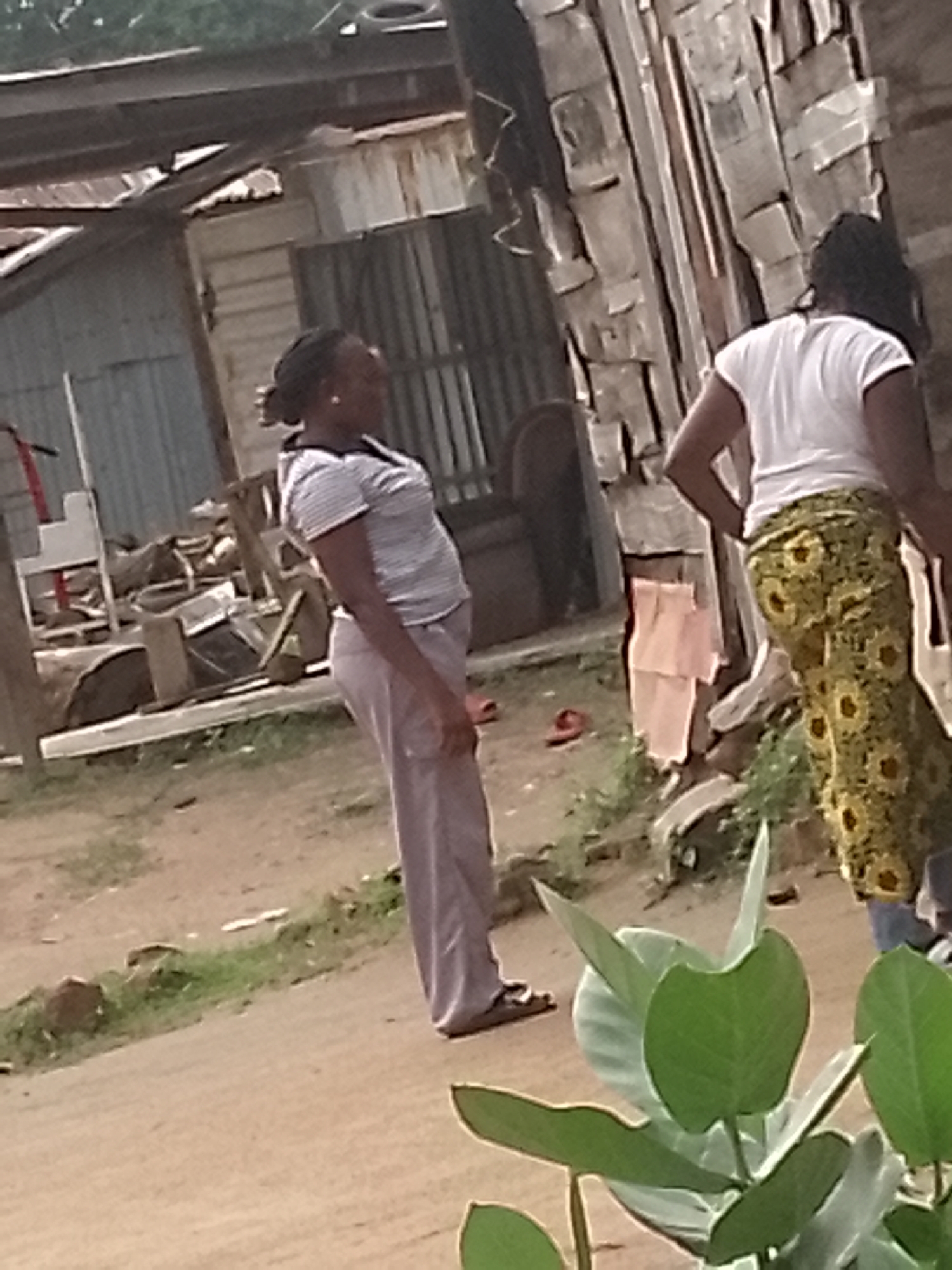 Two women stand in what appears to be a somewhat dilapidated outdoor area in Adankolo, Nigeria. The woman on the left, with her hair in a bun, wears a horizontally striped t-shirt and long, light-colored trousers. She is facing to her right, with her body slightly angled away from the camera, and her left hand rests on her hip. The woman on the right, facing away from the camera, has dark hair that falls down her back. She is wearing a white t-shirt and a long, yellow skirt with a distinctive floral or circular pattern. She has her left hand on her hip. In the background, there are structures that appear to be made of corrugated metal and wood, with some supporting beams visible. There are also some piles of debris and what looks like a broken chair. The ground is a mix of dirt and patchy grass. In the foreground, there are several large green leaves of a plant. A red sandal is visible on the ground near the woman on the left, and another red sandal is visible further in the background.