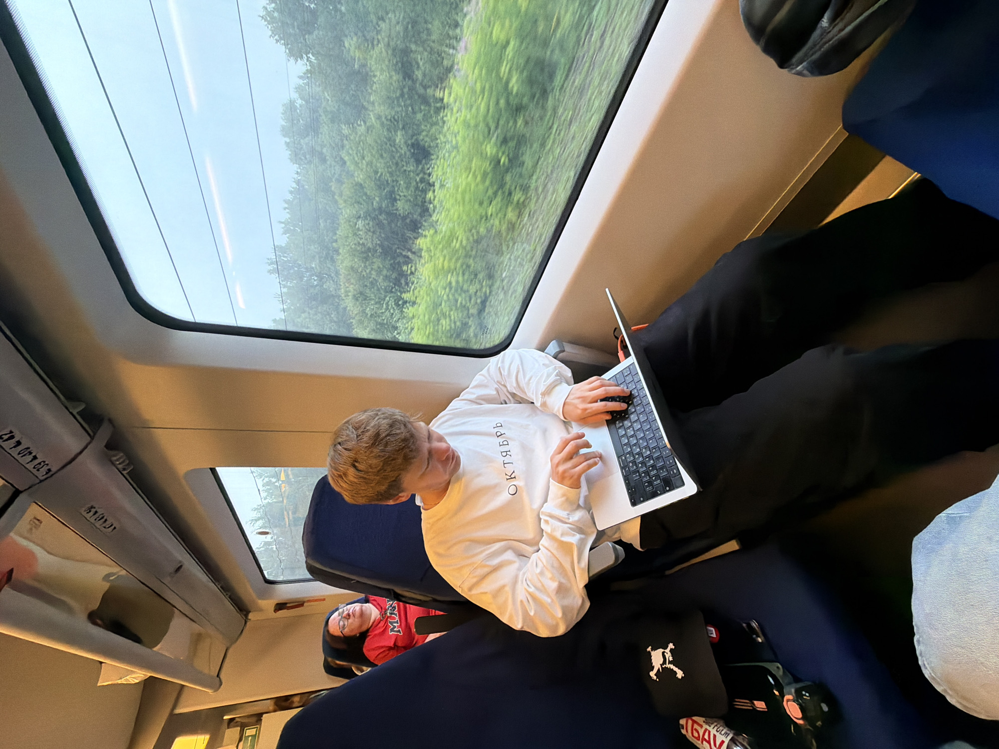 The image depicts the interior of a train carriage, likely in the city of Savostyanovo, Russia, given the text on the young man's sweater which reads "ОКТЯБРЬ" (October). A young man with light brown hair is seated in the foreground, engrossed in using a silver laptop placed on his lap. He is wearing a white long-sleeved sweatshirt with the aforementioned Russian text and black pants. To his left, a woman with dark hair and glasses is seated, wearing a red t-shirt, and appears to be sleeping or resting with her eyes closed. The train carriage features blue seats and a large window to the right, through which lush green trees and foliage are visible, suggesting the train is traveling through a rural or suburban area. Overhead compartments and part of the train's interior lighting are also visible. On the seat next to the young man, a black baseball cap with a white graphic and a bottle with a red and white label are placed. The overall lighting suggests it is daytime, possibly overcast or evening.