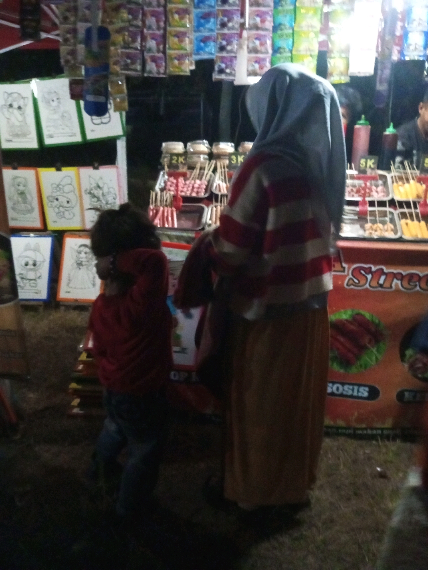 The image depicts a street scene at night, likely a marketplace or fair in Ngadikerso, Indonesia. The foreground is dominated by two individuals. On the right, a person wearing a grey hijab and a horizontally striped red and white long-sleeved shirt stands with their back to the camera, their lower body obscured by a long, tan skirt. To their left, a child in a red long-sleeved shirt and blue pants faces away from the camera, with their head tilted down.
Behind the individuals, a food stall is visible. It is adorned with colorful hanging items and displays of food. Several trays of what appear to be sausages or hot dogs on sticks are arranged, some labeled with prices like "2K" and "5K". A sign on the stall features the text "Street" in large letters, with smaller text below that is difficult to decipher but seems to relate to food.
In the background, more stalls are set up, with various items, including what look like coloring pages or art prints featuring cartoon characters, displayed on green and yellow backgrounds. The overall ambiance suggests a lively, outdoor event during the evening. The ground appears to be covered in dry grass or dirt, consistent with an outdoor setting.