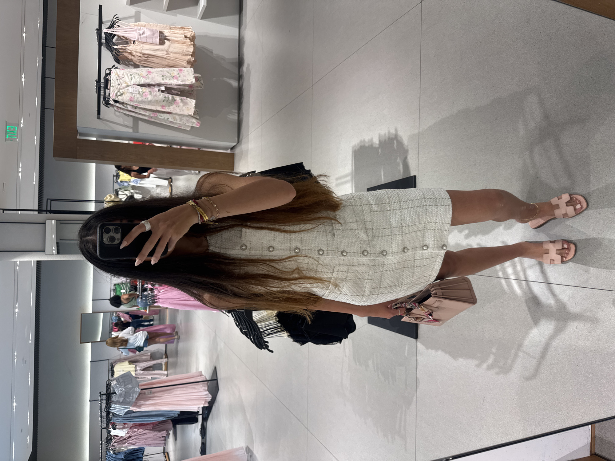 A young woman stands in a brightly lit clothing store, taking a mirror selfie. She is wearing a sleeveless, cream-colored tweed dress with silver buttons down the front. She holds a small, light pink handbag with a colorful scarf tied to its handle. On her feet are light pink slides. She has long, dark brown hair and wears several bracelets on her left wrist and a ring on her right index finger. The background shows racks of clothing, display shelves, and other shoppers in the distance. The floor is tiled. The setting is a retail environment, likely a fashion boutique or department store, potentially located in Miami, United States of America, given the overall aesthetic and common retail establishments in that area.