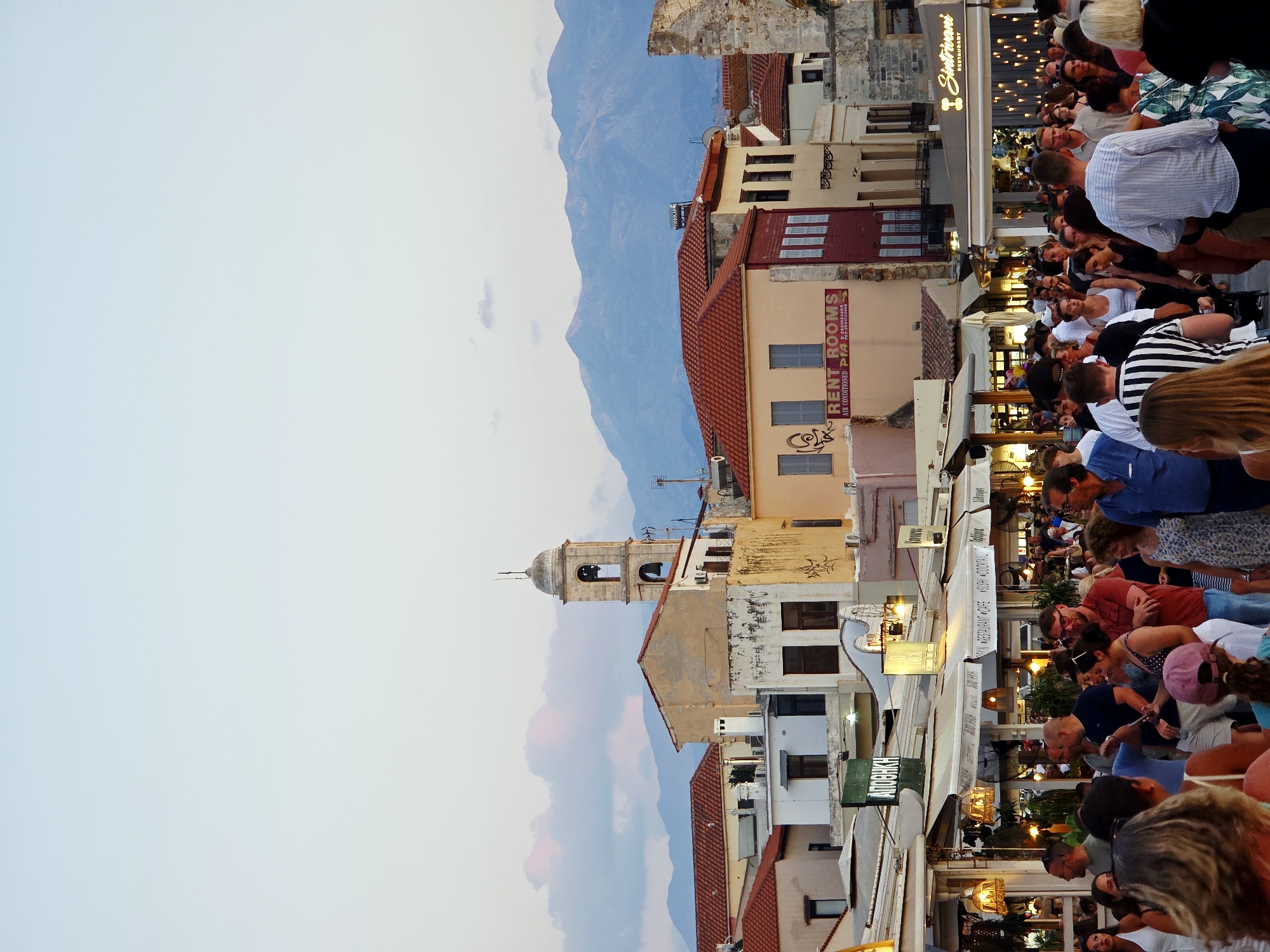The image depicts a bustling street scene in the old town of Chania, Greece, likely during the evening. In the background, characteristic Venetian-era buildings with red-tiled roofs dominate the skyline. A tall bell tower, a common feature in Chania's architecture, rises above the buildings. Further in the distance, the silhouette of the White Mountains is visible against a pastel-colored sky, tinged with pink and blue from the setting sun.
The foreground and midground are filled with a large crowd of people walking along a pedestrian street lined with shops and restaurants. The establishments feature awnings, outdoor seating, and illuminated signs, indicating they are open for business. Some signs are in Greek, such as "ΑΠΟΘΗΚΗ" (Apothiki, meaning warehouse or store) and "RENT ROOMS." The atmosphere appears lively, with people engaged in conversation and moving in various directions. The attire of the people suggests a warm climate, with many wearing casual summer clothing. The overall impression is one of a vibrant tourist destination, popular with both locals and visitors.