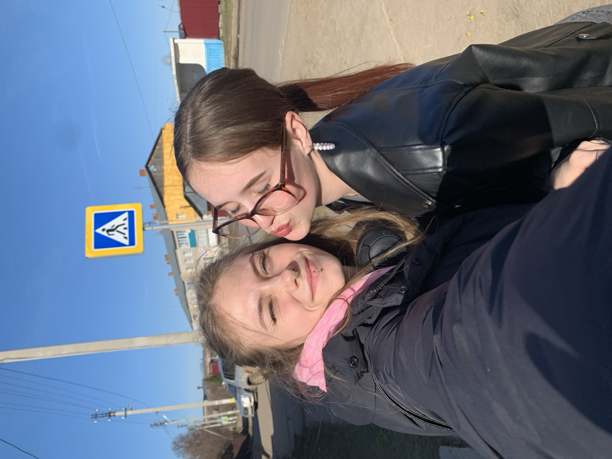 The image, taken in Foki, Russia, captures a playful outdoor selfie of two young women under a clear blue sky.
In the foreground, a young woman with light brown hair is smiling with her eyes partially closed, appearing to squint in the bright sunlight. She is wearing a black puffer jacket with a visible pink inner layer, possibly a scarf or shirt. She has several facial piercings, including an eyebrow ring, a septum ring, and labret piercings.
Behind her, a second young woman with darker hair, wearing glasses and a black leather jacket, leans in, making a puckering gesture towards the first woman. She has a distinctive white beaded ear cuff on her visible ear.
The background shows an urban or suburban setting with various buildings, including a yellow house and other structures, under a clear, bright blue sky. A blue and yellow pedestrian crossing sign is visible on the left, depicting a walking figure. Utility poles and power lines are also noticeable in the background. The bright sunlight indicates it is daytime. The overall mood conveyed by the subjects' expressions is happy and affectionate.