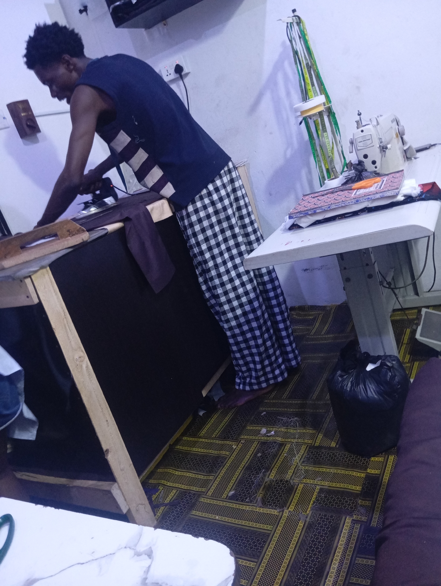 In this indoor shot, a young man is seen ironing clothes at a tailor's workbench. He is wearing a dark blue sleeveless shirt with horizontal stripes and a black and white checkered skirt. The workbench, made of wood and covered with a dark fabric, is cluttered with fabric and ironing tools. A sewing machine sits on a table next to the workbench, with spools of thread and measuring tapes hanging on a stand. The floor is covered with a patterned rug. A black trash bag is placed on the floor near the table. The lighting suggests it might be evening or an indoor setting with artificial light. The overall scene depicts a busy tailoring workshop.