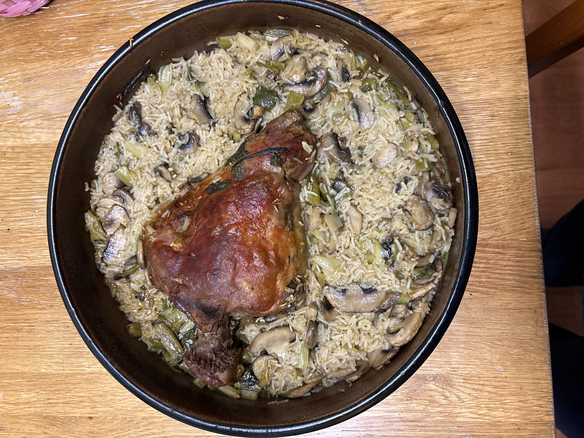 A round, black, ceramic dish filled with rice, mushrooms, and leeks, with a roasted lamb shank on top, sits on a wooden table. The photo appears to have been taken from directly above. The location is likely somewhere in the United Kingdom, possibly Oswestry. No people are visible in the image.