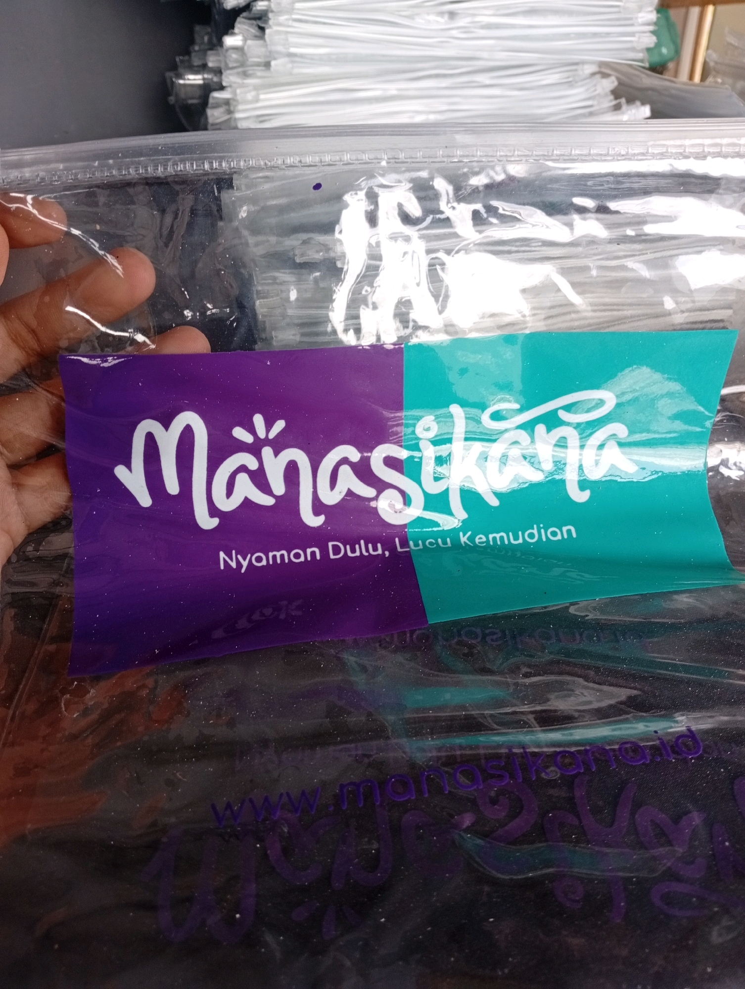 The image shows a close-up of a clear plastic bag containing a purple and teal colored label. The label has the text "Manasikana" written in white cursive font, with "Nyaman Dulu, Lucu Kemudian" written below it in smaller white font. The bag is transparent, allowing a dark, possibly fabric, item to be seen underneath. The background of the image shows stacks of white, plastic-wrapped items, potentially more bags or products. A hand is visible on the left side of the image, holding the plastic bag open. The lighting is bright, causing reflections on the plastic. The location is unknown.